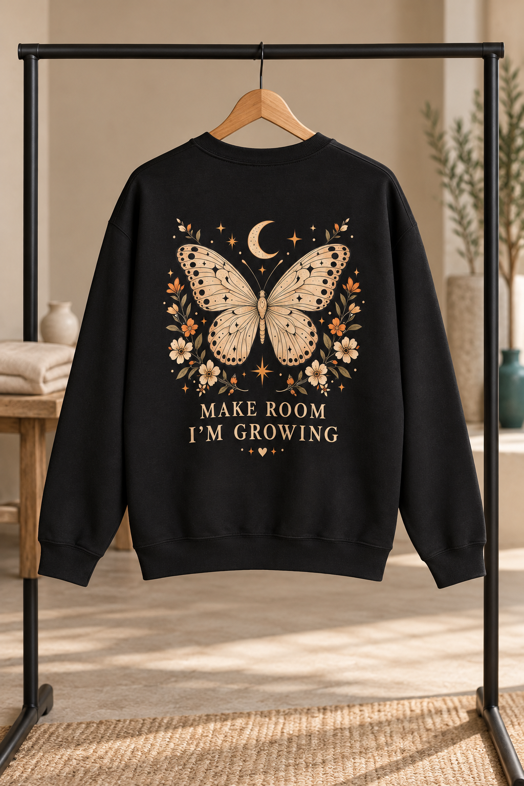 A black crewneck sweatshirt hanging on a wooden hanger from a black clothing rack, featuring a large graphic of a cream-colored butterfly with floral details and the text 'MAKE ROOM I'M GROWING' in cream lettering.