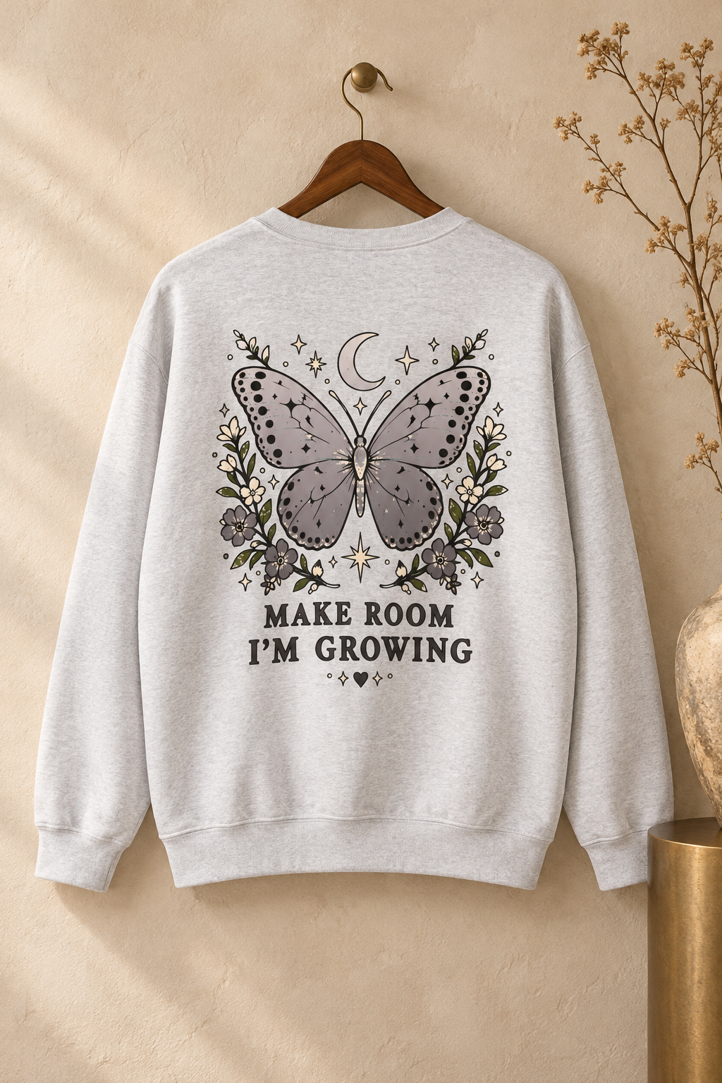 A light heather gray crewneck sweatshirt hanging on a wooden hanger against a warm beige wall. The back of the sweatshirt features a graphic of a butterfly surrounded by flowers, a crescent moon, and stars, with the text 'MAKE ROOM I'M GROWING' printed below.