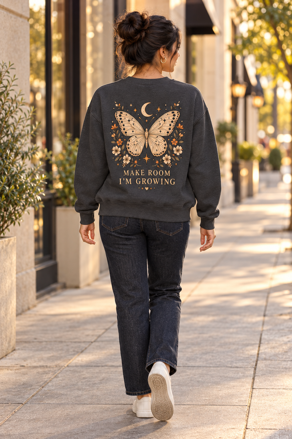 A woman viewed from behind walking on a sunlit city sidewalk, wearing a dark charcoal sweatshirt with a large butterfly, moon, stars, and floral graphic on the back, plus the text 'MAKE ROOM I'M GROWING.' She is also wearing dark jeans and white sneakers.