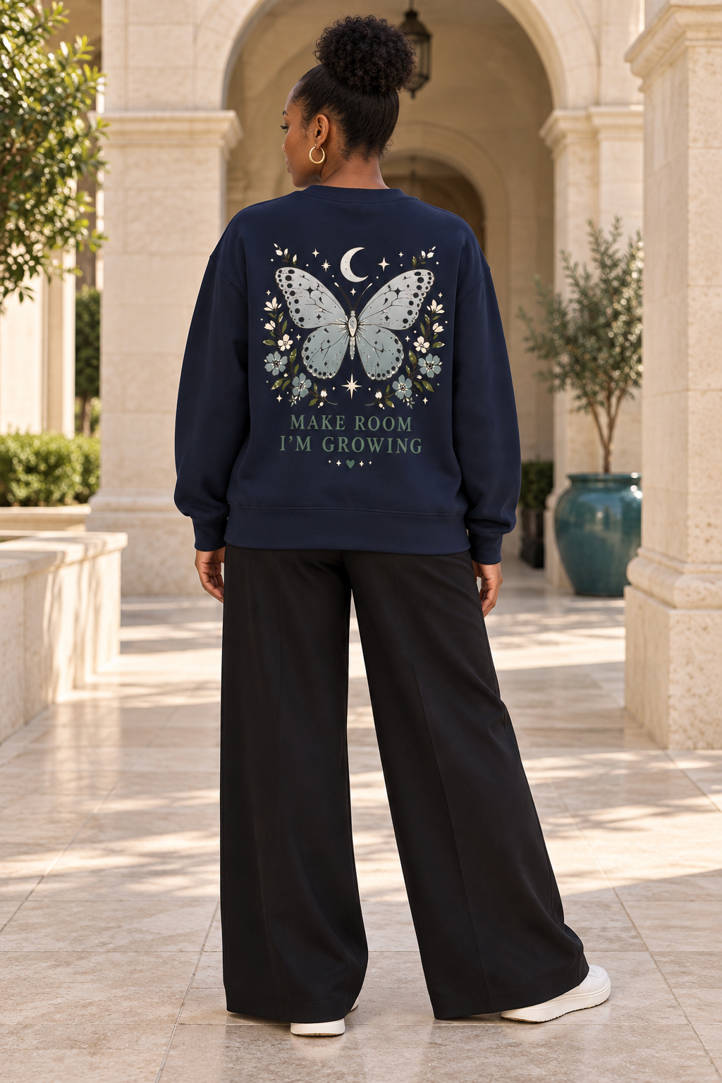 A woman viewed from behind wearing a navy blue sweatshirt with a large graphic on the back featuring a butterfly, stars, a crescent moon, and the text 'MAKE ROOM I'M GROWING'. She is also wearing black wide-leg pants and white sneakers, standing in a sunlit outdoor corridor with stone columns and potted plants.