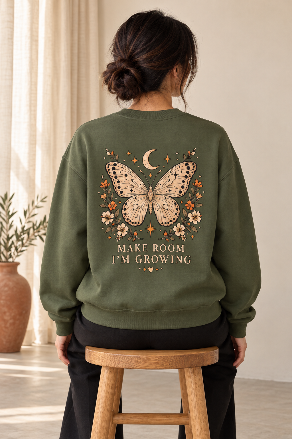 A woman viewed from behind wearing an olive green sweatshirt with a large butterfly, moon, flowers, and the text 'MAKE ROOM I'M GROWING' printed on the back. She is seated on a wooden stool in a softly lit indoor room.