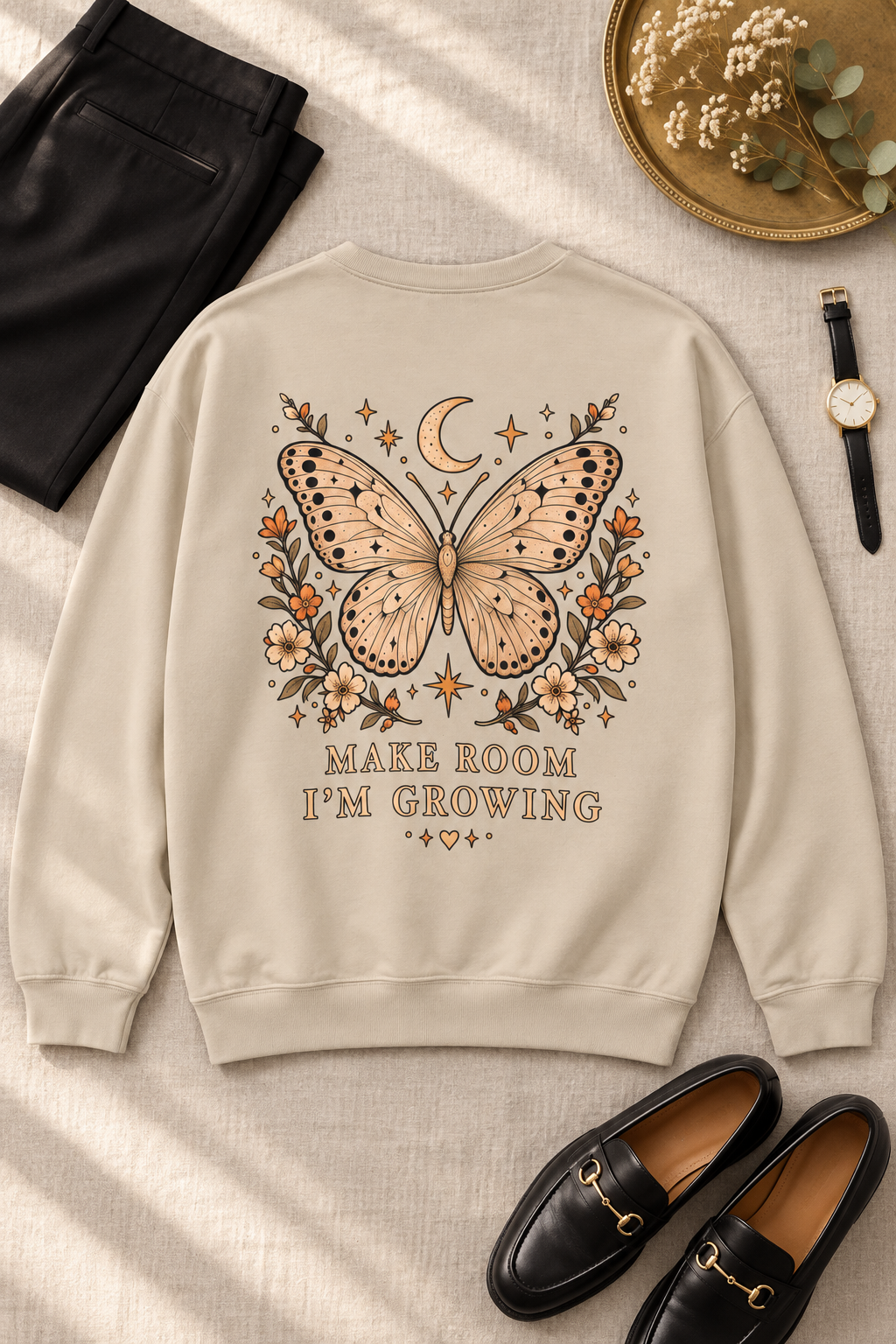 A beige sweatshirt with a large butterfly, moon, and floral graphic on the back, styled in a flat lay with black pants, black loafers, a watch, and a gold tray on a textured beige fabric background.
