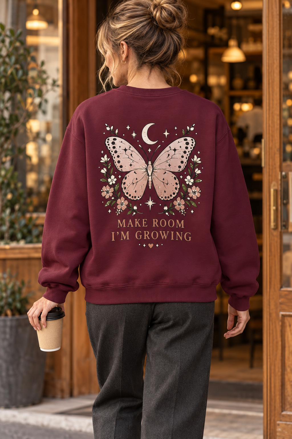 A woman viewed from behind wearing a maroon crewneck sweatshirt with a large butterfly, moon, stars, and floral graphic on the back, above the text 'MAKE ROOM I'M GROWING'. She is holding a takeaway coffee cup in a shop doorway.