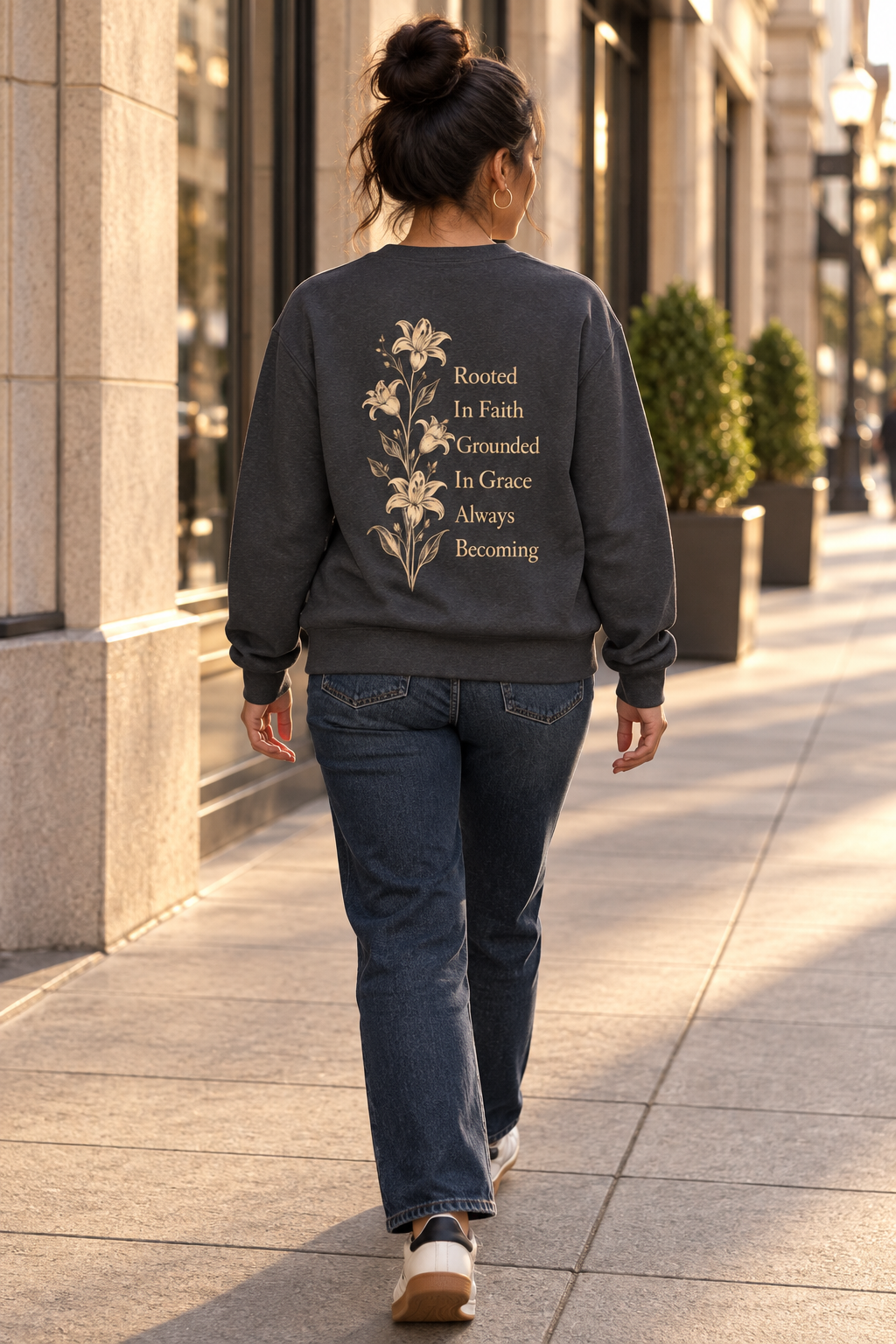 A woman viewed from behind walking on a city sidewalk, wearing a dark charcoal gray crewneck sweatshirt with a floral and inspirational quote graphic on the back, paired with blue jeans.