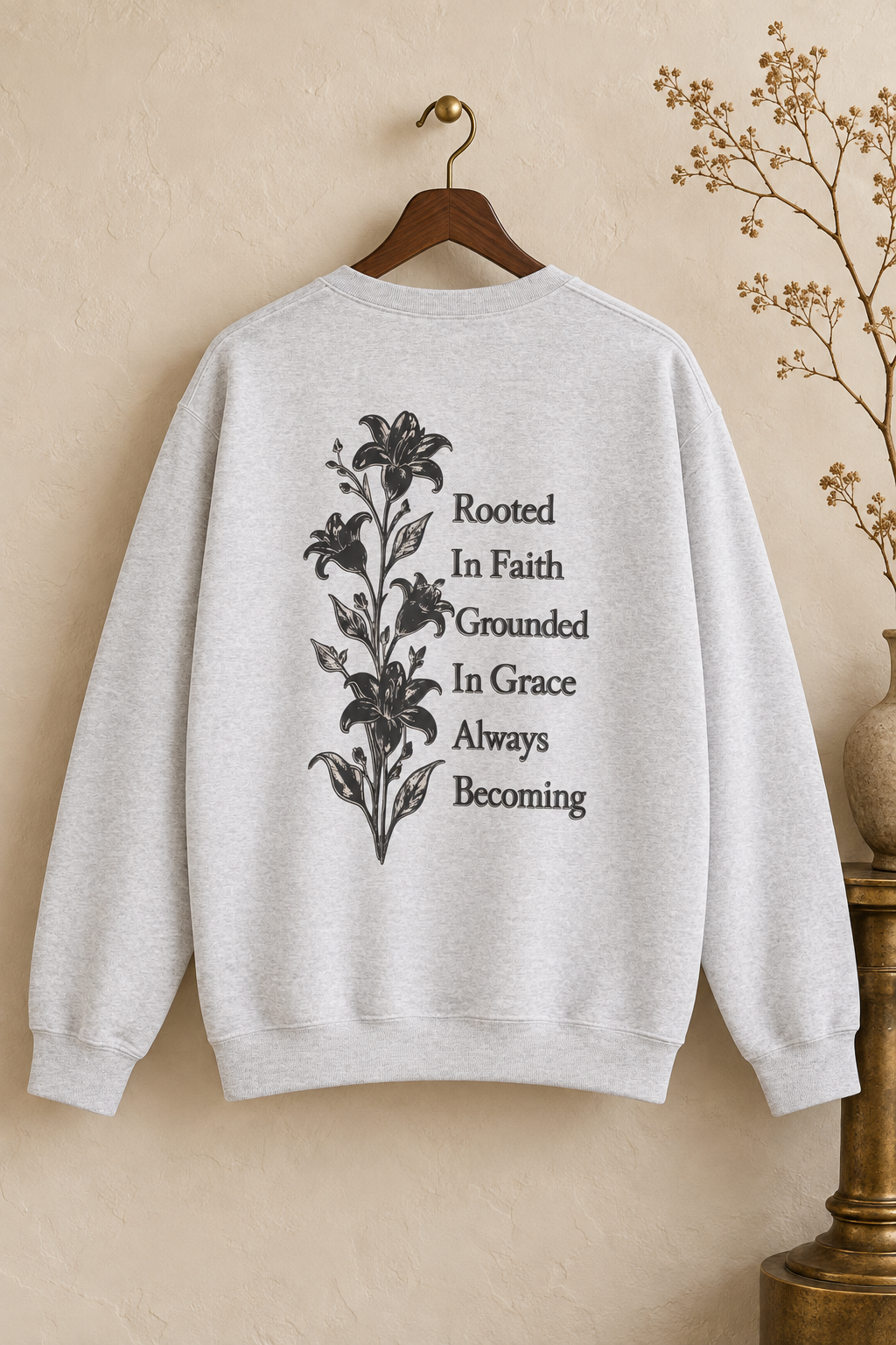 A light heather gray crewneck sweatshirt hanging on a wooden hanger against a textured beige wall. The back of the sweatshirt features a black graphic of lilies next to the text 'Rooted In Faith Grounded In Grace Always Becoming'.
