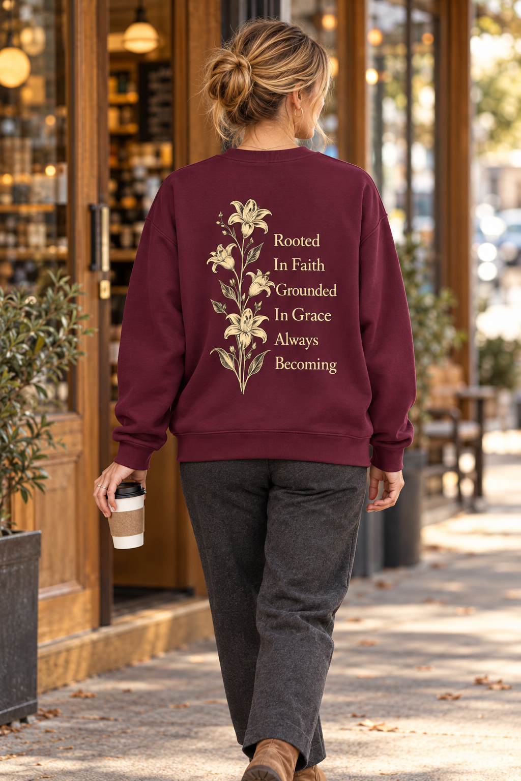 A woman viewed from behind walking on a sidewalk, wearing a burgundy crewneck sweatshirt with a cream floral and text graphic on the back reading 'Rooted In Faith Grounded In Grace Always Becoming'.