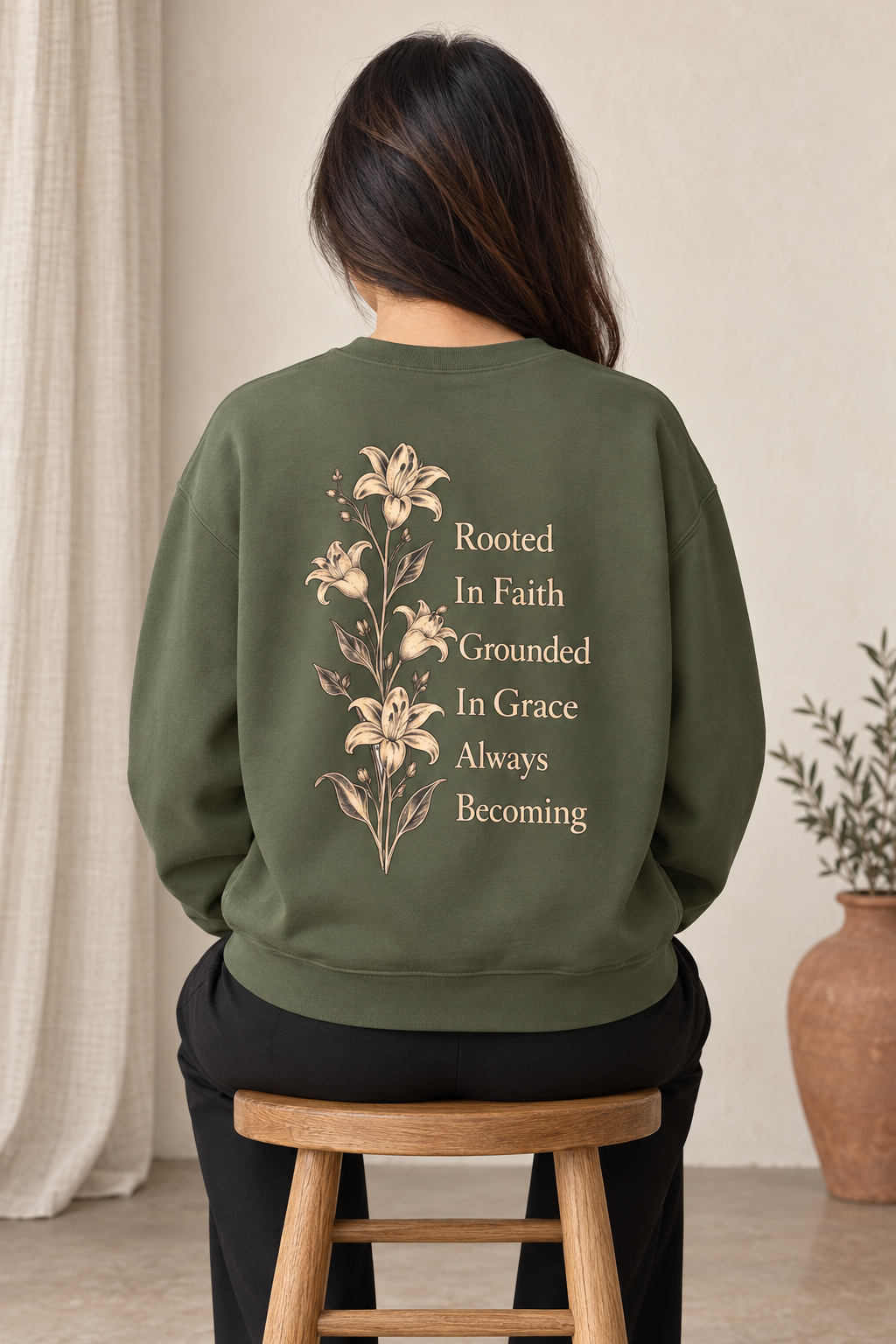 A woman viewed from behind wearing an olive green sweatshirt with a beige floral and text graphic on the back, sitting on a wooden stool in a neutral indoor setting.
