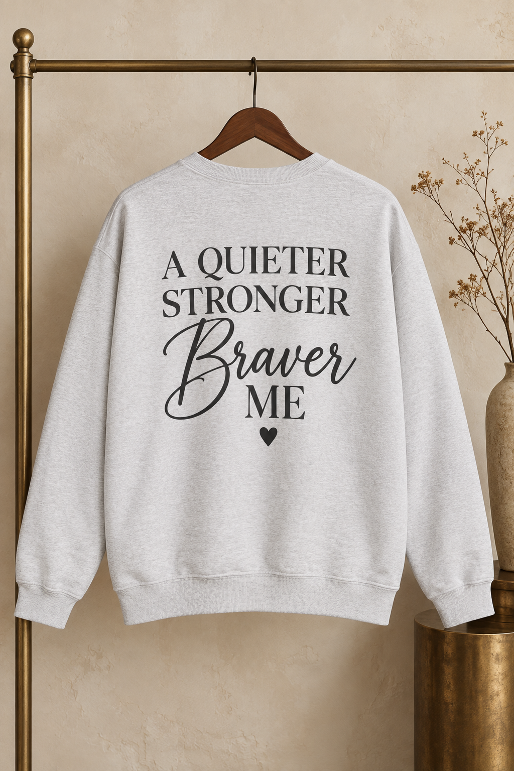 A light heather gray crewneck sweatshirt hanging on a wooden hanger from a gold clothing rack, featuring the text 'A QUIETER STRONGER Braver ME' in black lettering on the back.