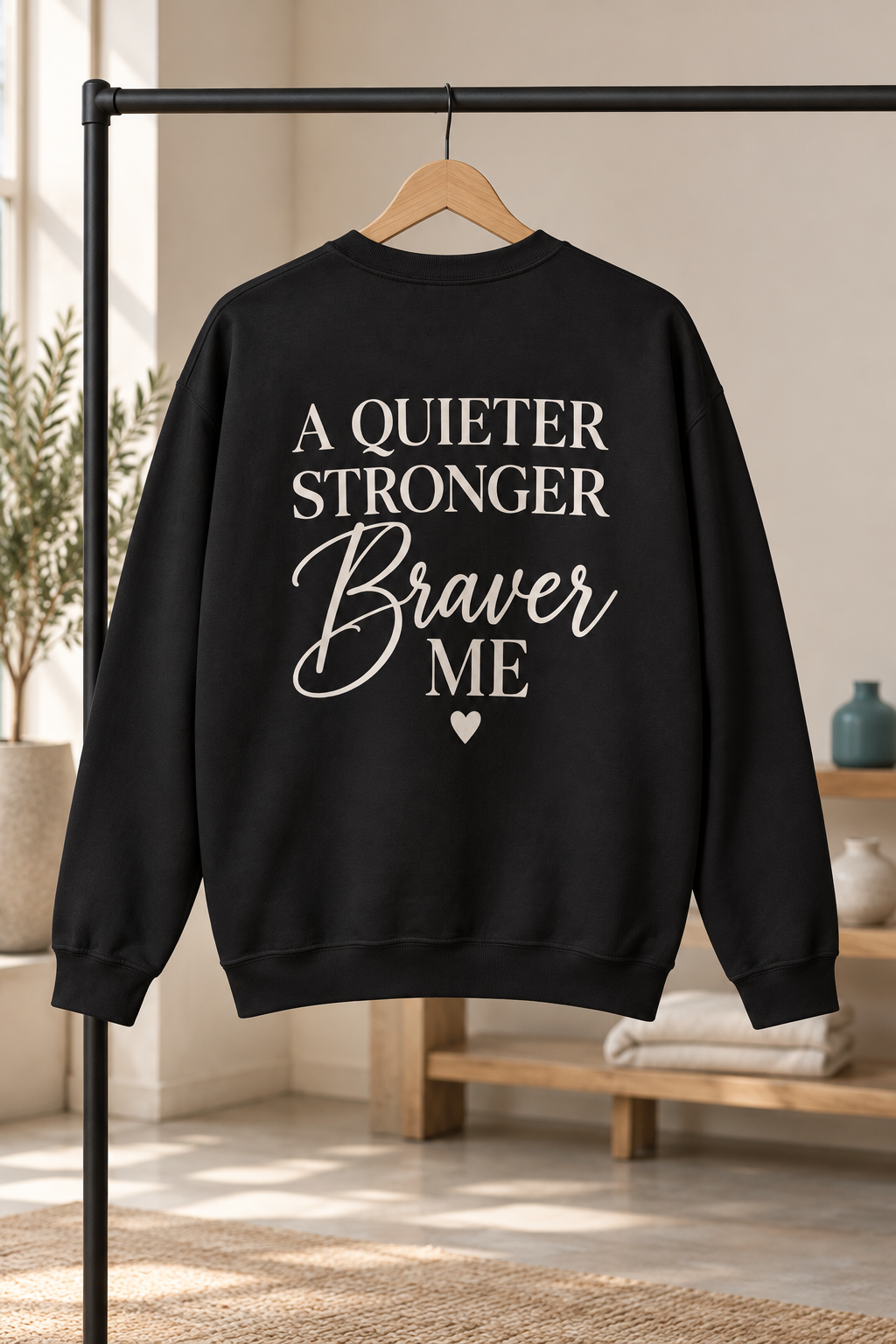 A black crewneck sweatshirt hanging on a wooden hanger from a black clothing rack, featuring the phrase 'A QUIETER STRONGER Braver ME' in cream text on the back, set against a softly blurred warm-toned interior background.