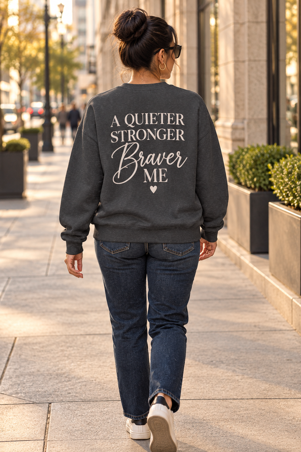 A woman viewed from behind walking on a city sidewalk, wearing a dark charcoal gray sweatshirt with the cream text 'A QUIETER STRONGER Braver ME' on the back, paired with dark blue jeans and white sneakers.