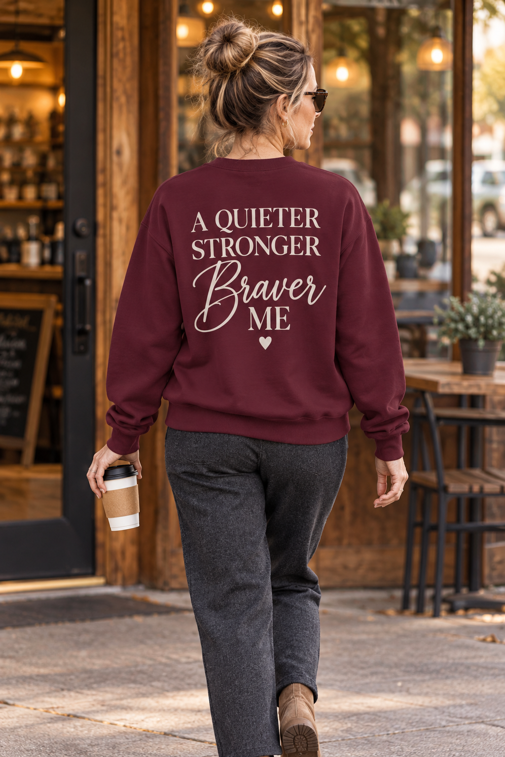 A woman seen from behind walking on a sidewalk in front of a coffee shop, wearing a maroon sweatshirt with the text 'A QUIETER STRONGER Braver ME' printed in cream on the back.