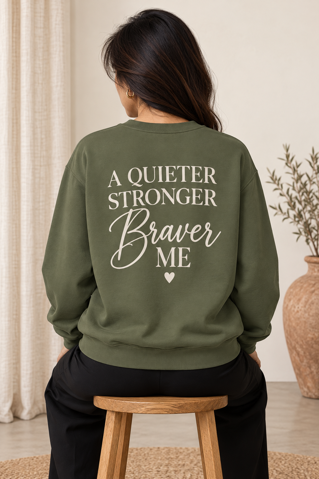 A woman viewed from behind wearing an olive green sweatshirt with the text 'A QUIETER STRONGER Braver ME' printed in cream across the back, seated on a wooden stool in a softly lit indoor setting.