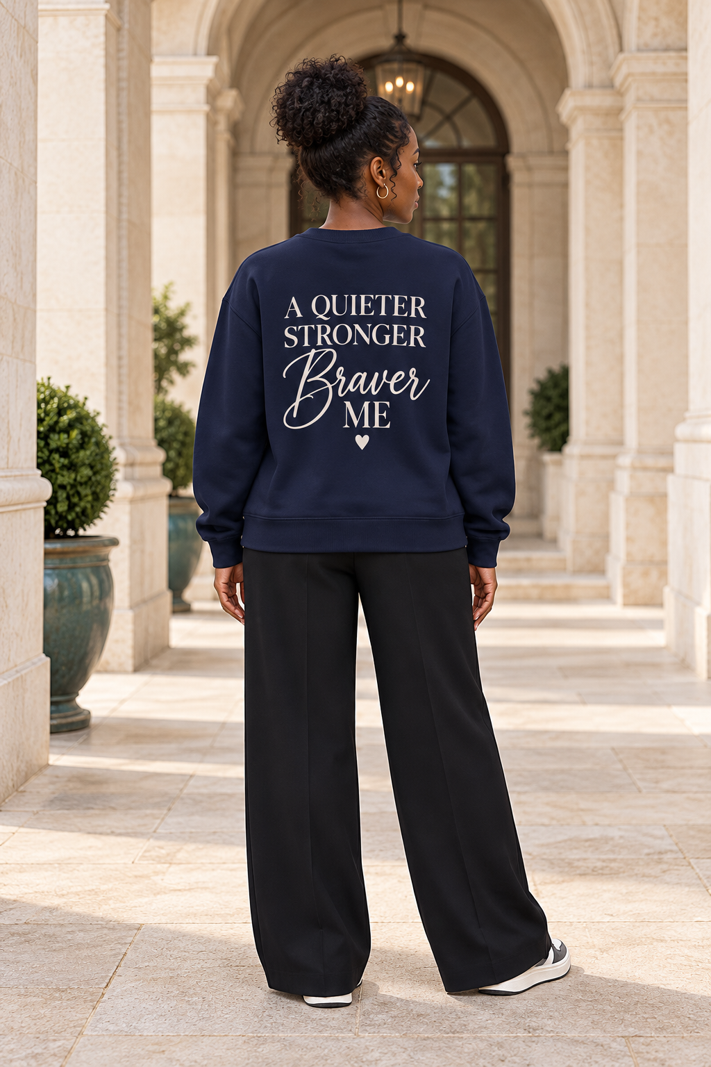 A woman viewed from behind wearing a navy blue sweatshirt with cream text that reads 'A QUIETER STRONGER Braver ME' and black wide-leg pants, standing in a stone colonnade.
