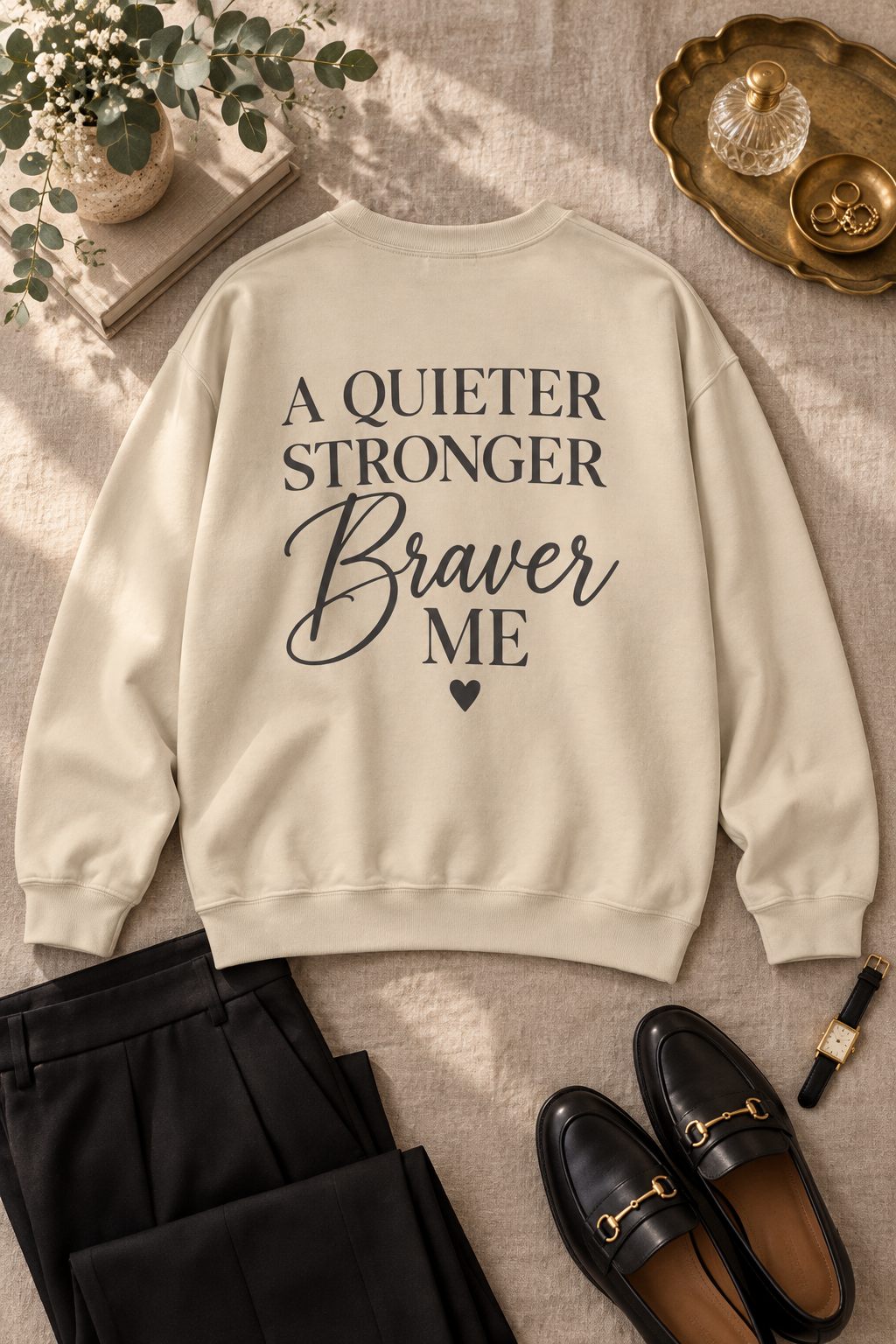 A beige crewneck sweatshirt with the text 'A QUIETER STRONGER Braver ME' printed in black on the front, styled in a flat lay with black trousers, black loafers, and a small potted plant on a textured beige fabric background.