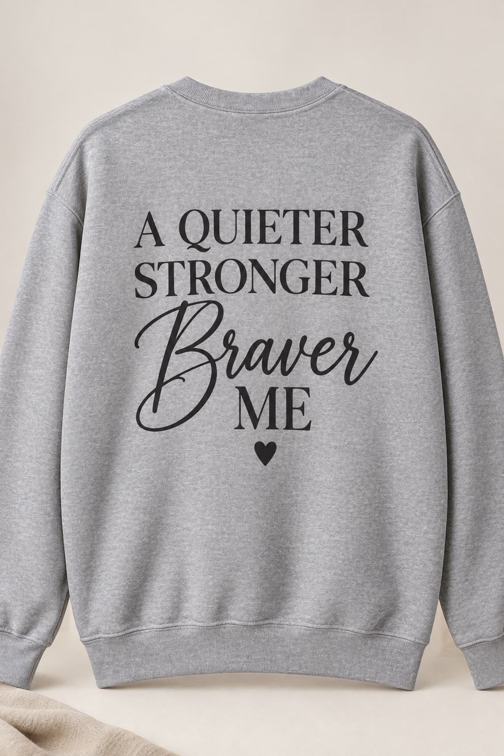 A heather gray crewneck sweatshirt shown from the back, featuring the text 'A QUIETER STRONGER Braver ME' in black lettering with a small heart below, set against a neutral beige background.