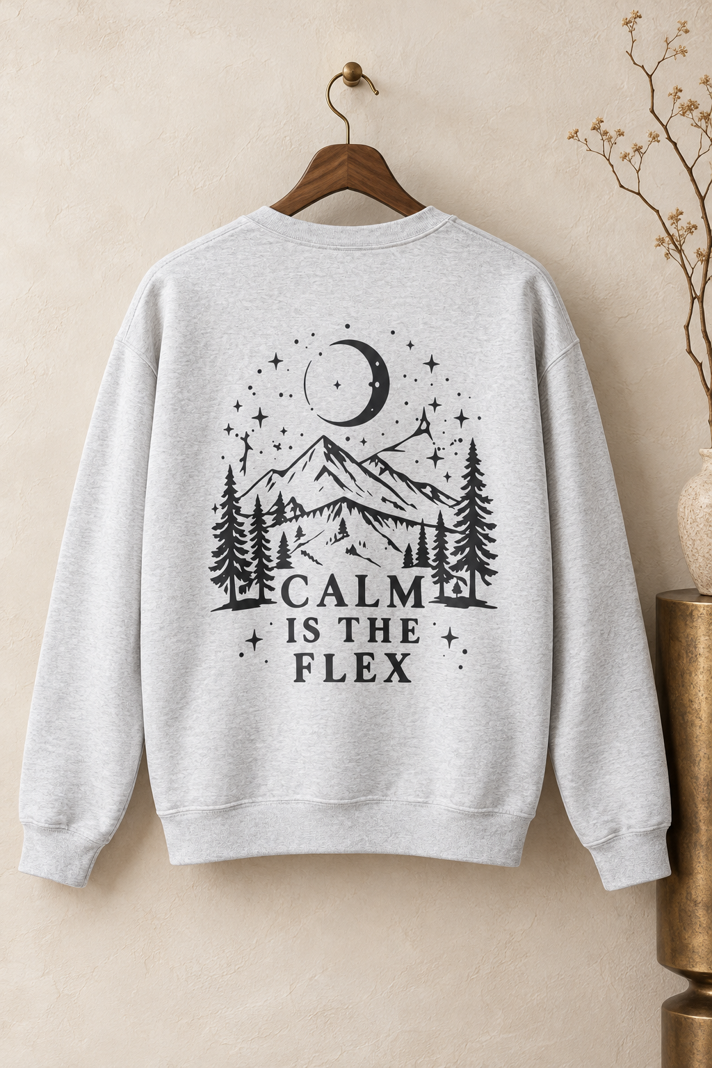 A light heather gray crewneck sweatshirt hanging on a wooden hanger against a textured beige wall. The back of the sweatshirt features a black graphic of a mountain, pine trees, a crescent moon, and stars, with the text 'CALM IS THE FLEX'.