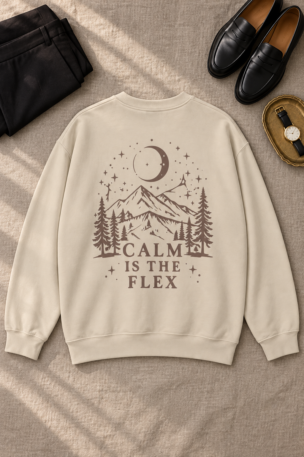 A beige crewneck sweatshirt with a brown graphic print of a mountain, pine trees, a crescent moon, and stars, with the text 'CALM IS THE FLEX'. The sweatshirt is displayed in a flat-lay arrangement on a textured beige fabric background, surrounded by black loafers, a black jacket, and a watch.