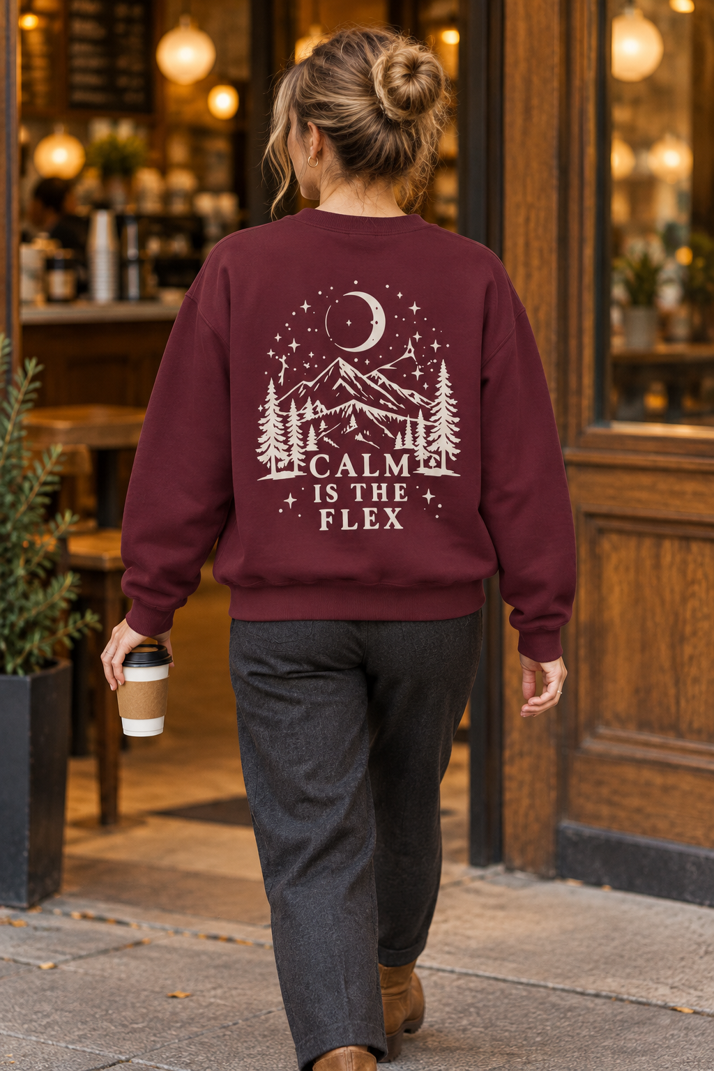 A woman viewed from behind walking out of a cozy cafe, wearing a maroon crewneck sweatshirt with a cream graphic of a mountain, trees, moon, and stars with the text 'CALM IS THE FLEX'.