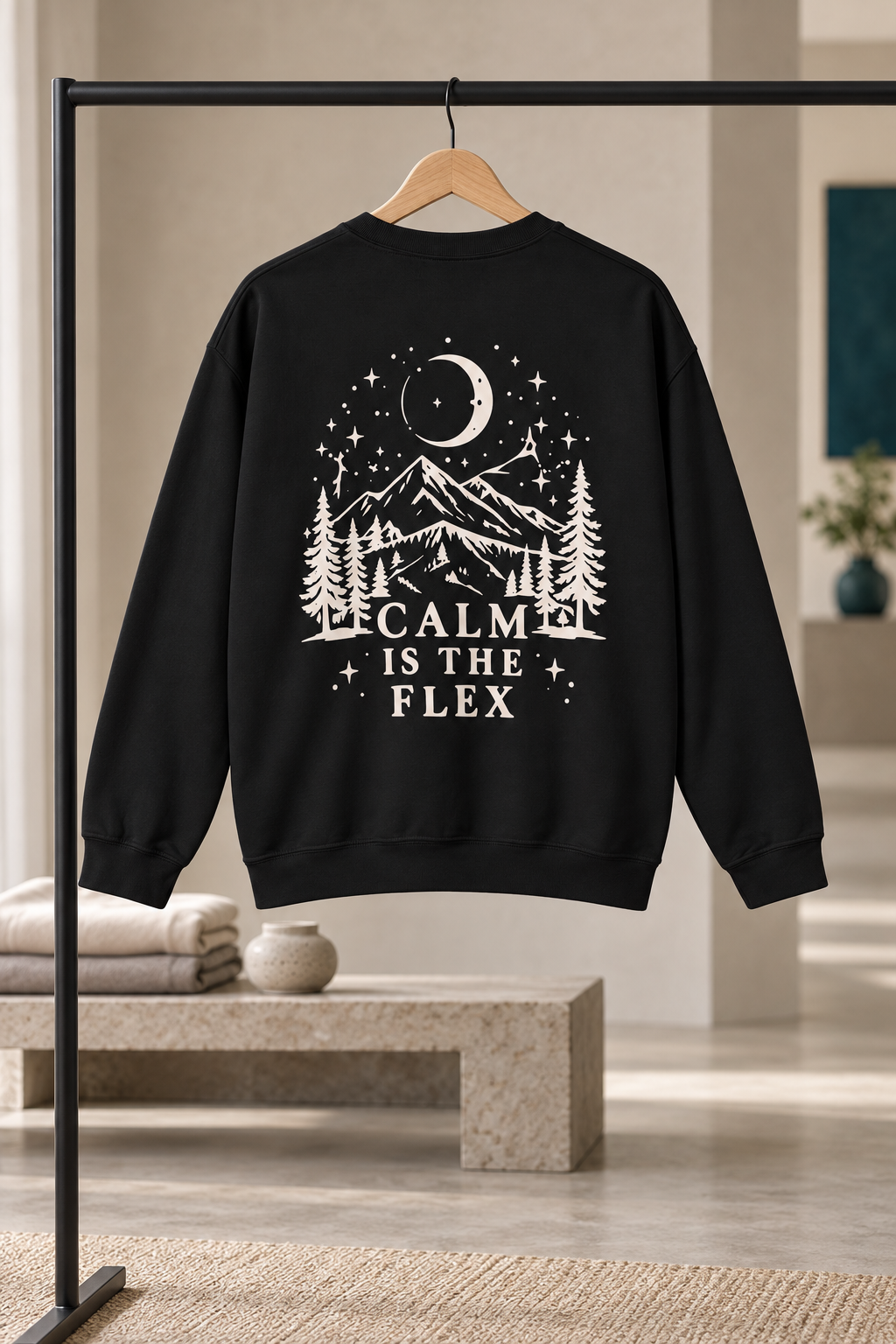 A black crewneck sweatshirt hanging on a wooden hanger from a black clothing rack, featuring a cream graphic print of a mountain, moon, stars, and pine trees with the text 'CALM IS THE FLEX'. The background is a softly blurred, modern interior room.
