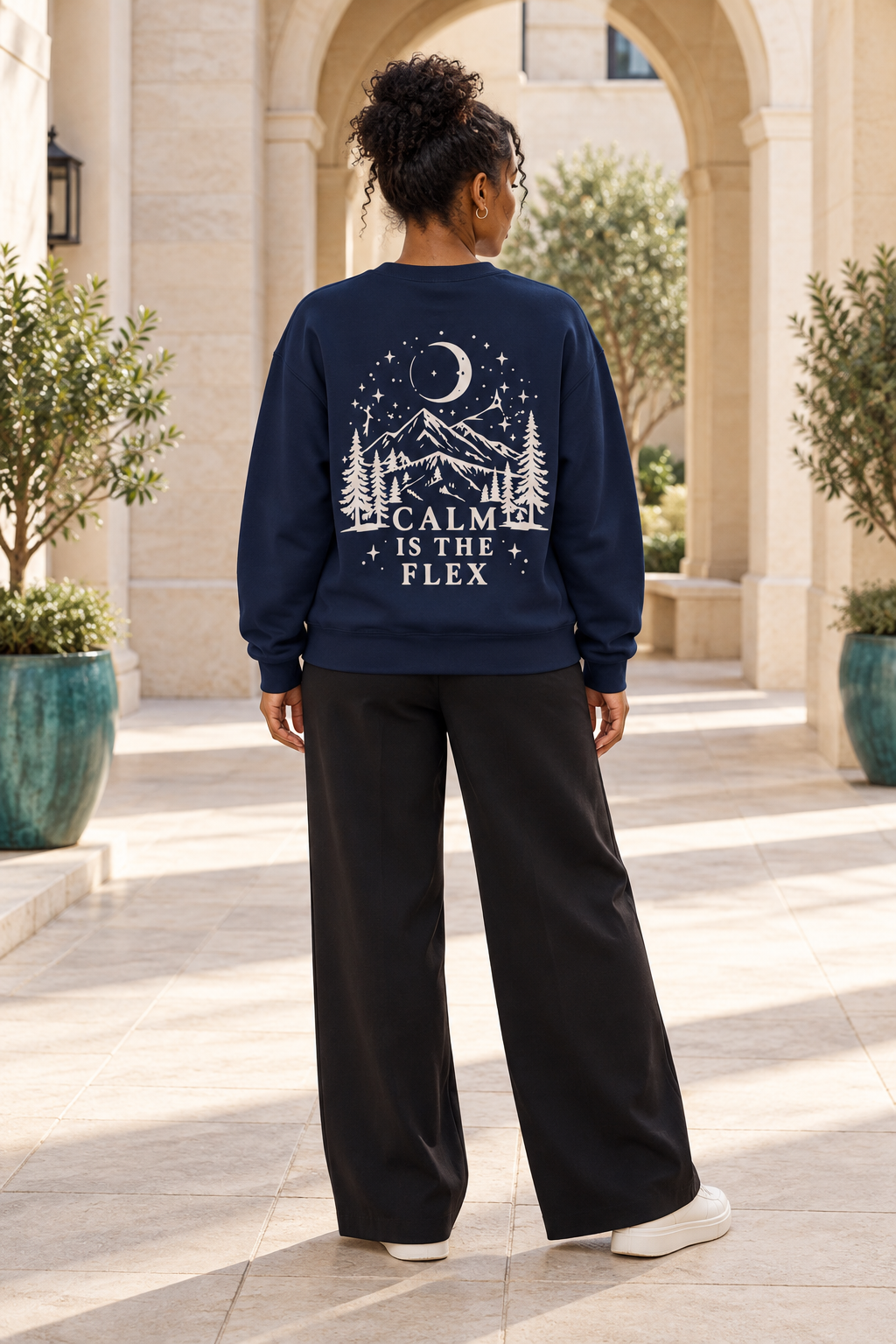 A woman viewed from behind wearing a navy blue crewneck sweatshirt with a cream graphic print of a mountain, trees, and stars with the text 'CALM IS THE FLEX'. She is also wearing black wide-leg sweatpants and white sneakers, standing in a sunlit outdoor walkway with stone arches and potted plants.