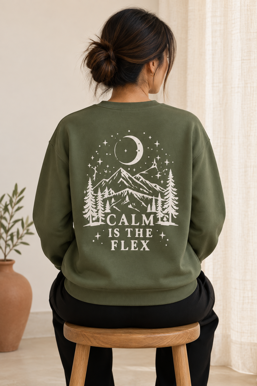 A woman viewed from behind wearing an olive green sweatshirt with a cream graphic print of a mountain, trees, moon, and stars, with the text 'CALM IS THE FLEX'. She is sitting on a wooden stool in a softly lit indoor setting.