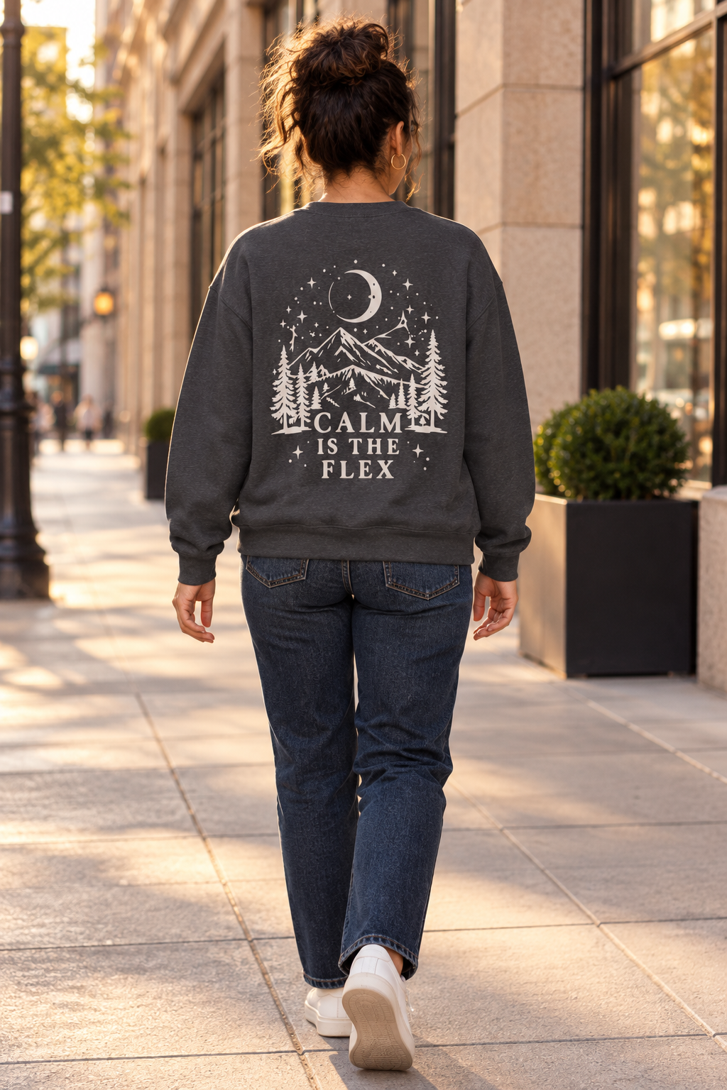A woman viewed from behind walking on a city sidewalk, wearing a dark charcoal gray crewneck sweatshirt with a white graphic of a mountain, trees, moon, and stars, and the text 'CALM IS THE FLEX'.