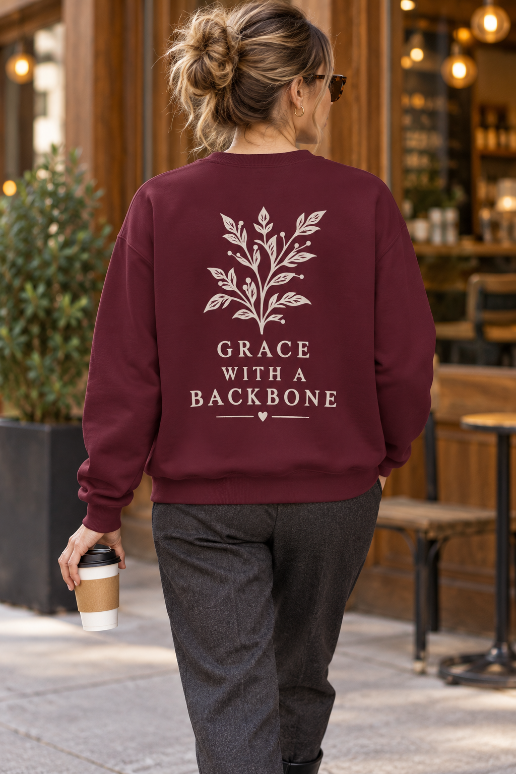 A woman viewed from behind wearing a maroon crewneck sweatshirt with a cream botanical leaf design and the text 'GRACE WITH A BACKBONE' printed on the back. She is walking outdoors in front of a cafe, holding a coffee cup.