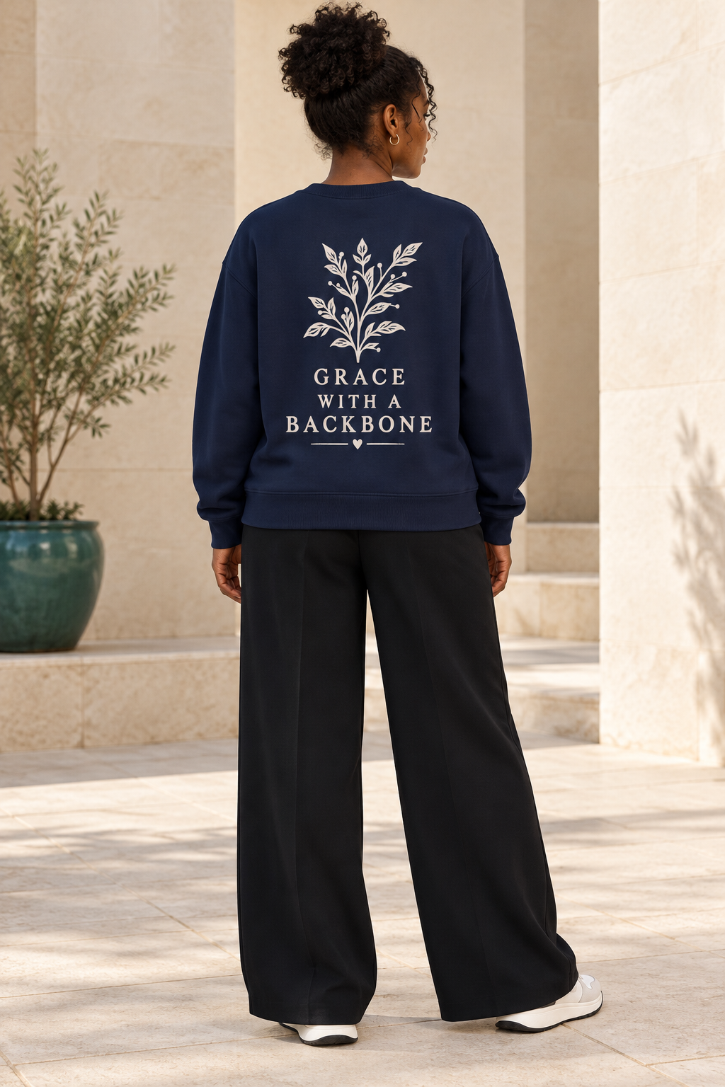 A woman viewed from behind wearing a navy blue sweatshirt with a cream graphic of a leafy branch and the text 'GRACE WITH A BACKBONE' on the back, paired with black wide-leg pants.