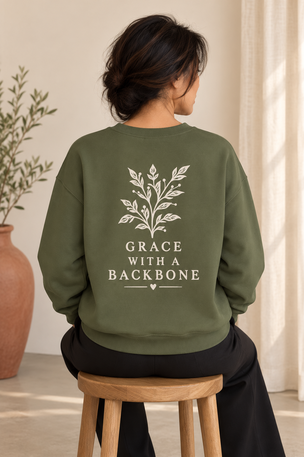 A woman viewed from behind wearing an olive green sweatshirt with a cream botanical leaf graphic and the text 'GRACE WITH A BACKBONE' printed on the back. She is seated on a wooden stool in a softly lit indoor setting with a potted plant and curtains in the background.
