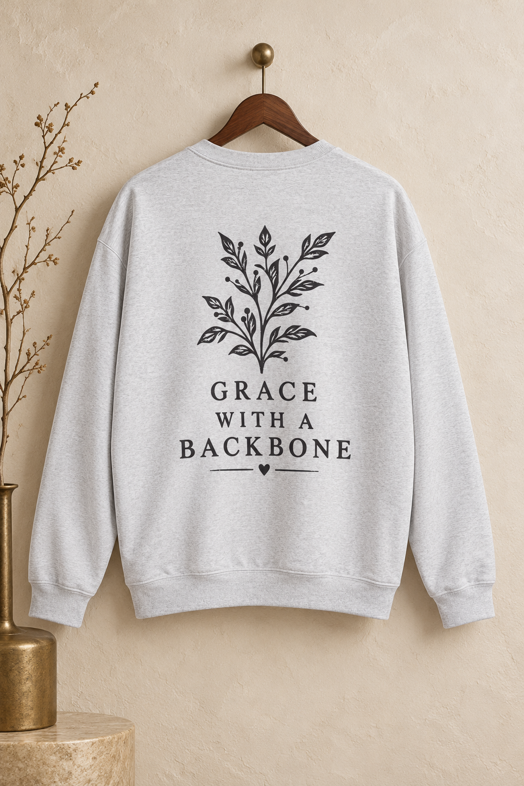 A light heather gray crewneck sweatshirt hanging on a wooden hanger against a textured beige wall. The back of the sweatshirt features a black graphic of a leafy branch with the text 'GRACE WITH A BACKBONE' and a small heart.
