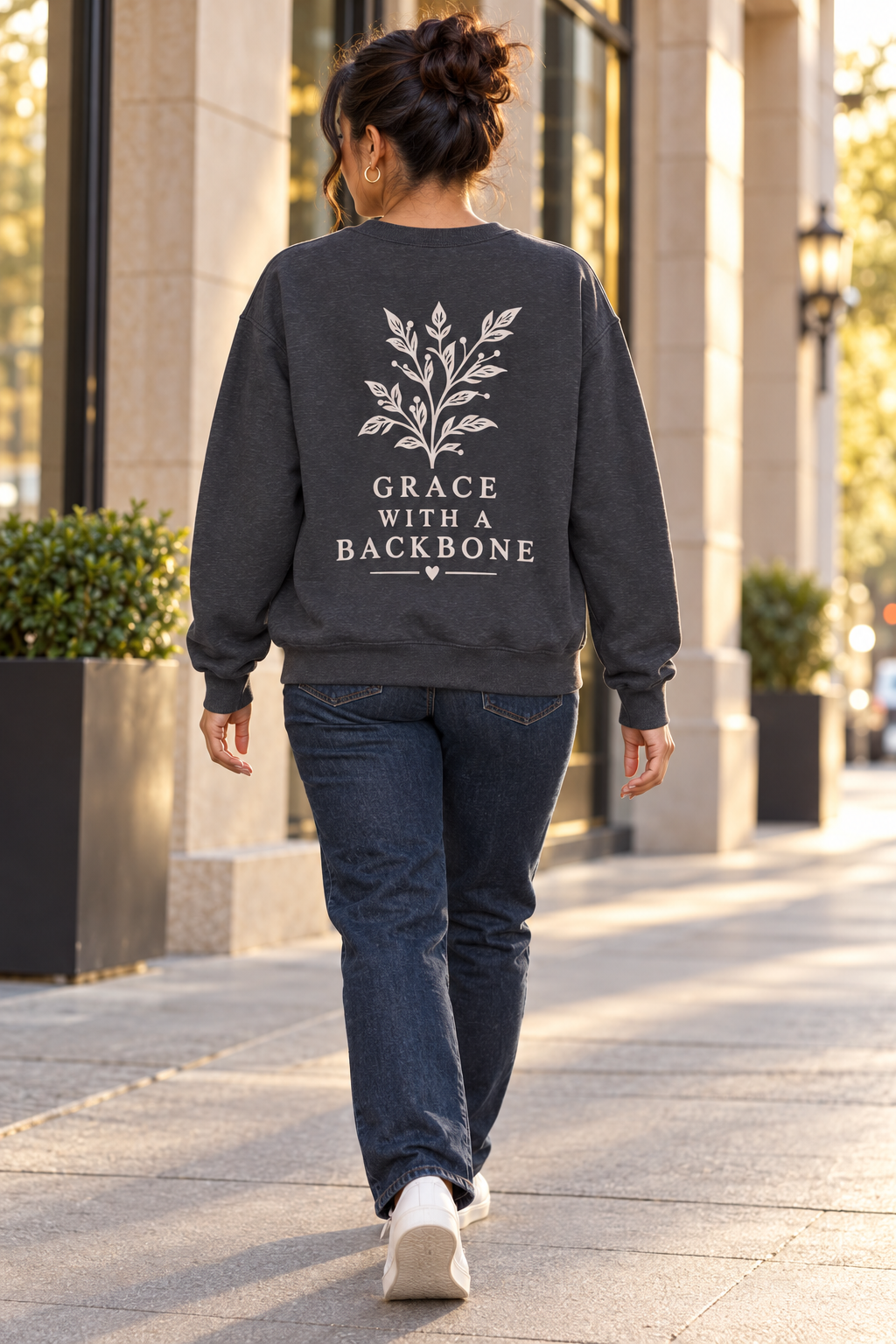 A woman viewed from behind walking on a city sidewalk, wearing a dark charcoal gray sweatshirt with a cream leaf graphic and the text 'GRACE WITH A BACKBONE' printed on the back, paired with dark blue jeans and white sneakers. 