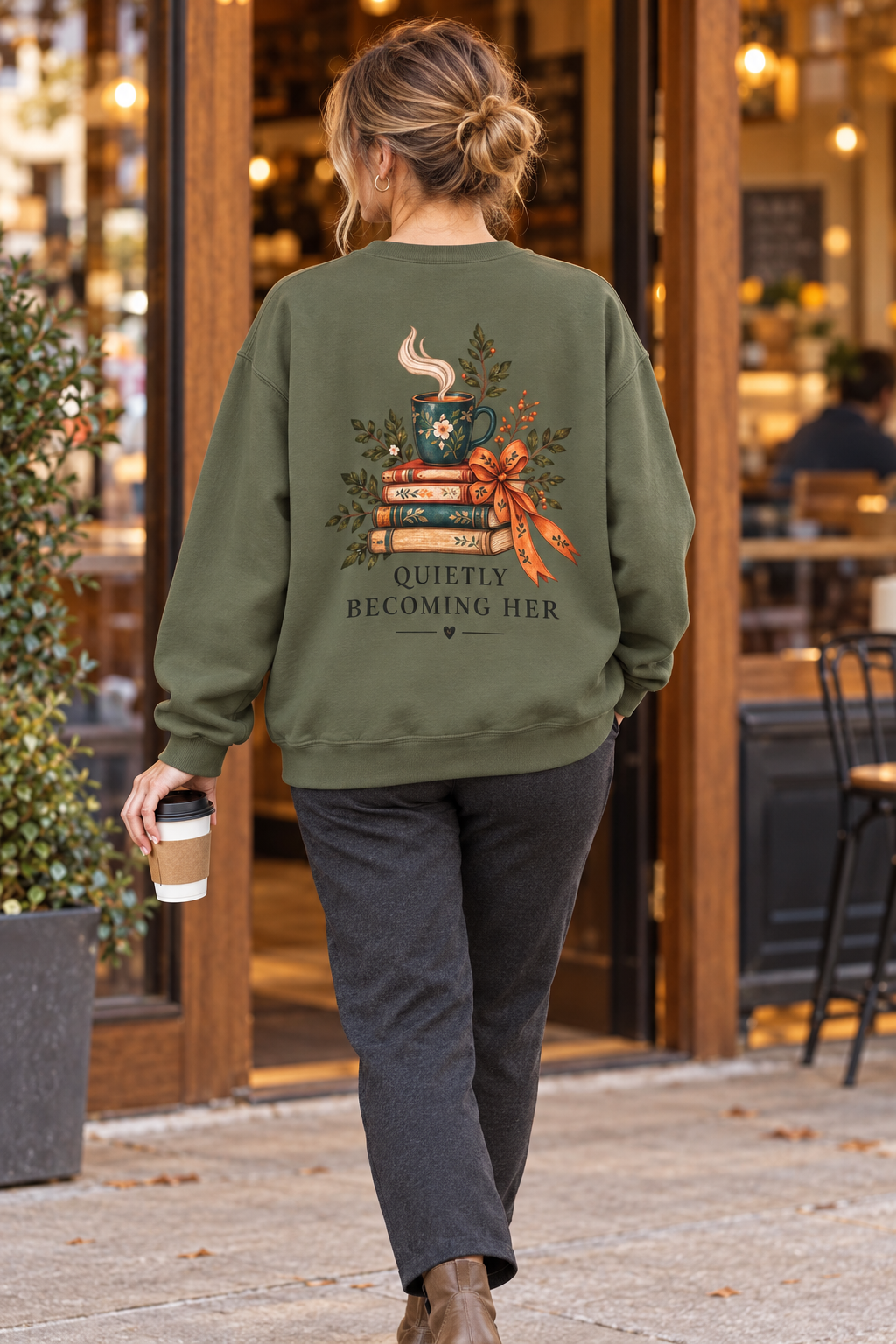 A woman viewed from behind walking outdoors in front of a cafe, wearing an olive green sweatshirt with a large graphic on the back featuring a book, ribbon, and the text 'QUIETLY BECOMING HER'.