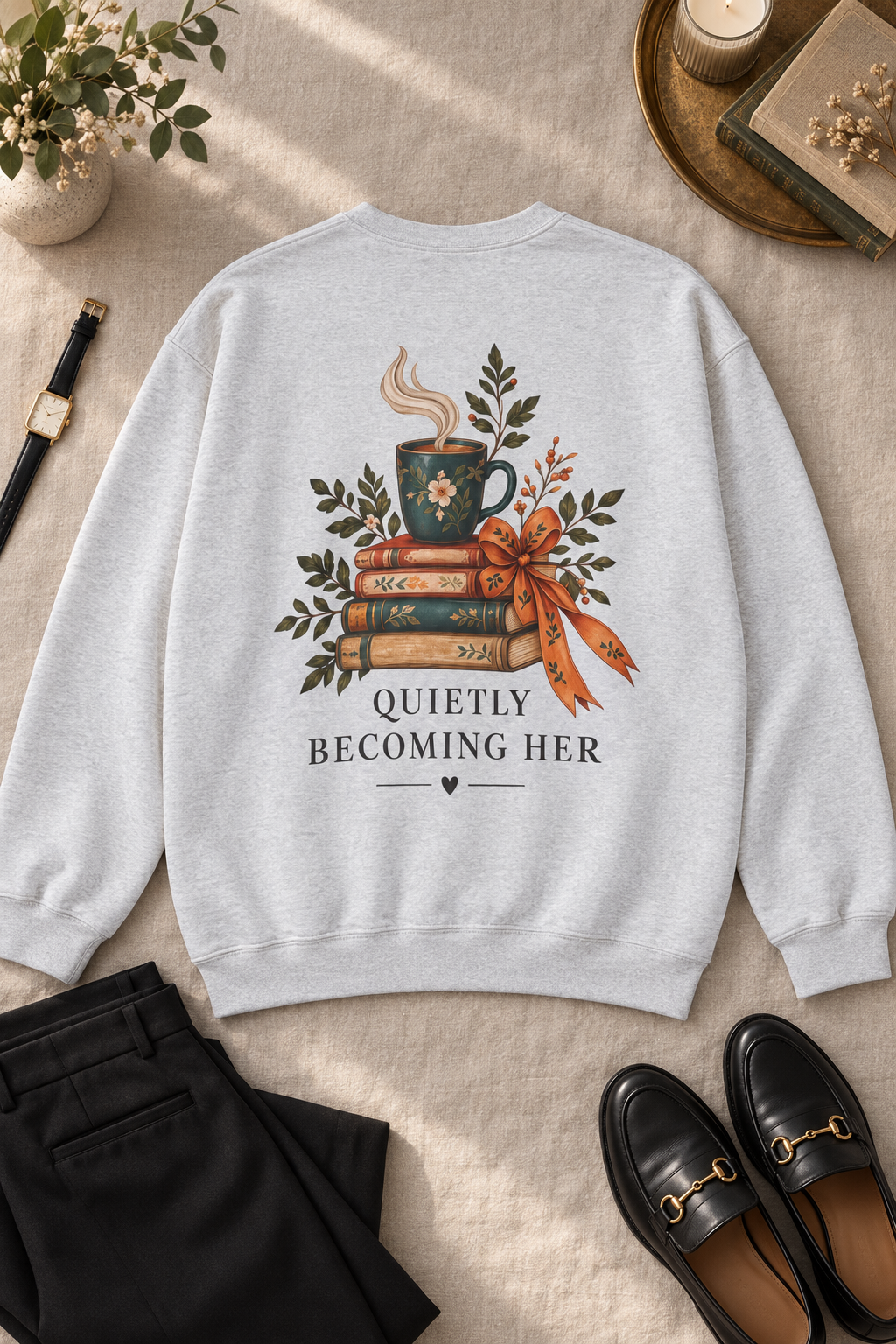 A light gray crewneck sweatshirt laid flat on a beige fabric surface, featuring a graphic of stacked books, a steaming mug, leaves, and the text 'QUIETLY BECOMING HER'. Black pants and black loafers are placed around it in a styled flat-lay arrangement with small decorative props.