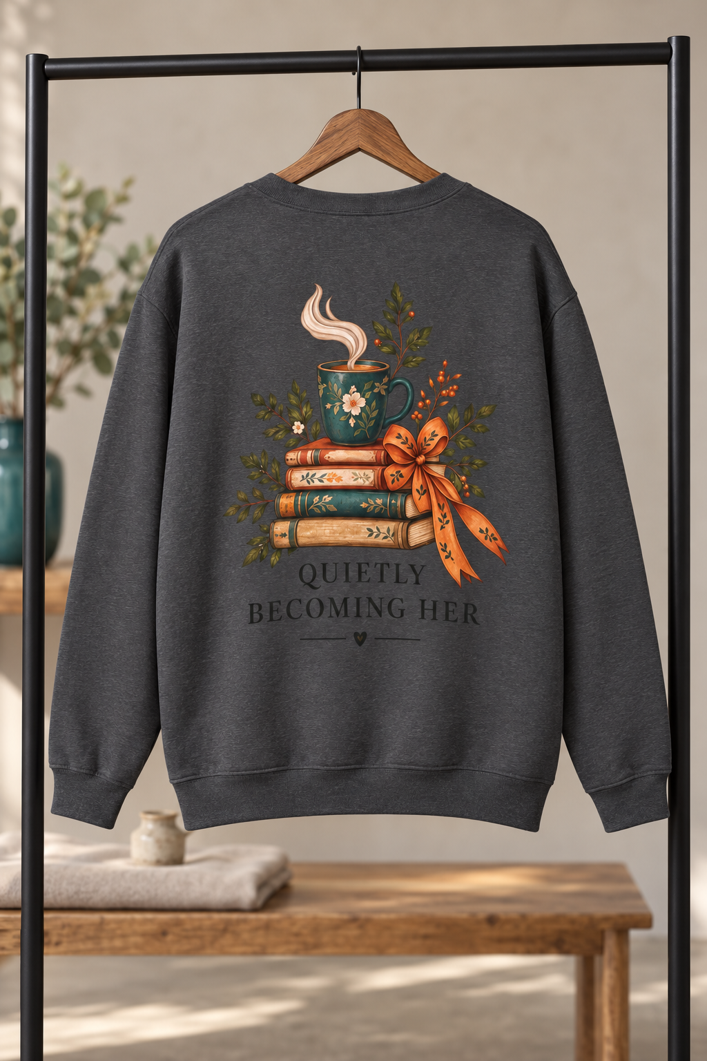 A dark gray crewneck sweatshirt hanging on a wooden hanger from a black clothing rack. The back of the sweatshirt features a graphic of a steaming teal mug resting on a stack of books, tied with a ribbon, with the text 'QUIETLY BECOMING HER' underneath.