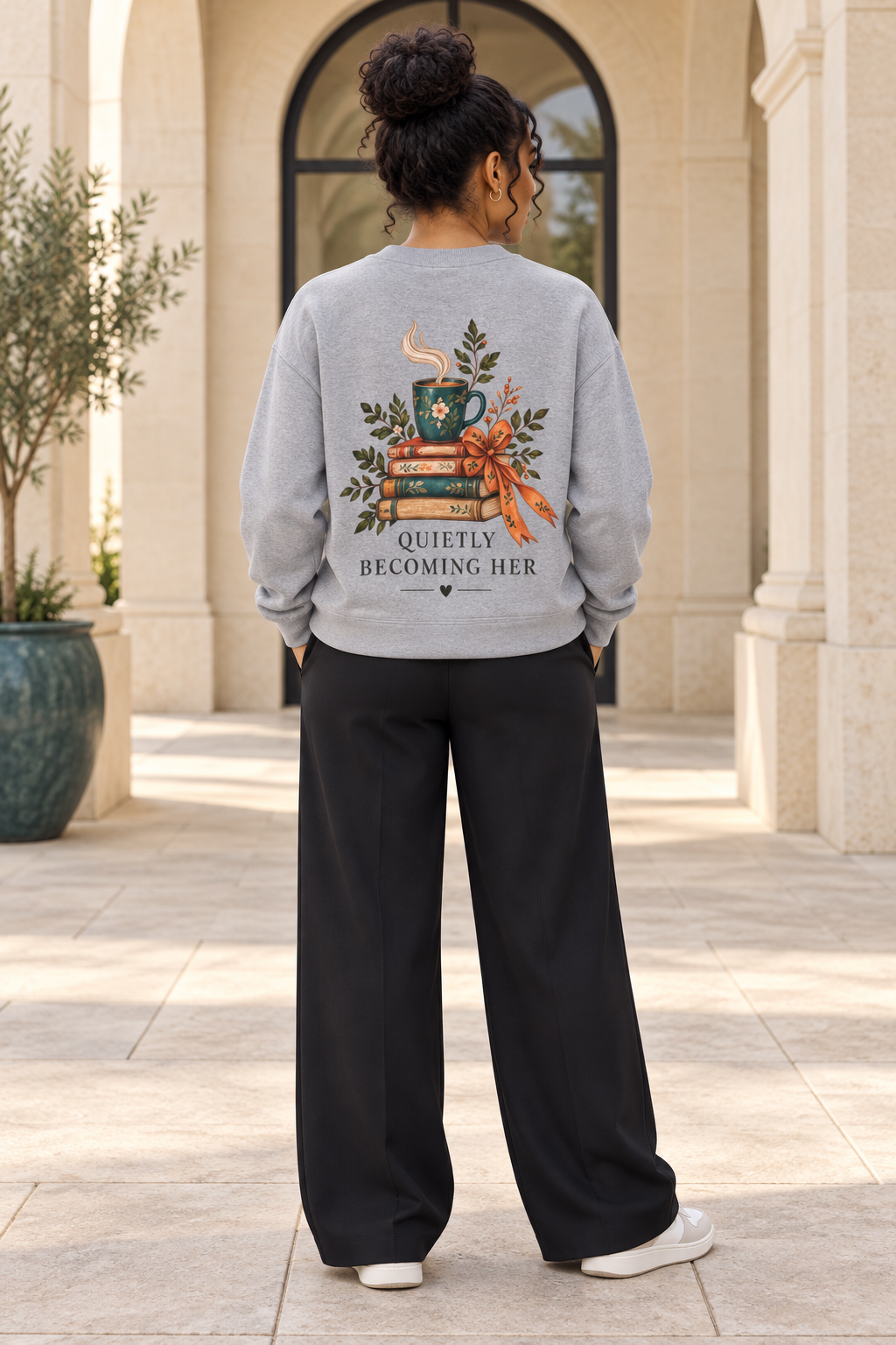 A woman viewed from behind wearing a light gray crewneck sweatshirt with a large graphic of stacked books, a coffee mug, and autumn leaves with the text 'QUIETLY BECOMING HER' on the back, paired with black wide-leg pants and white sneakers, standing in front of a stone building.