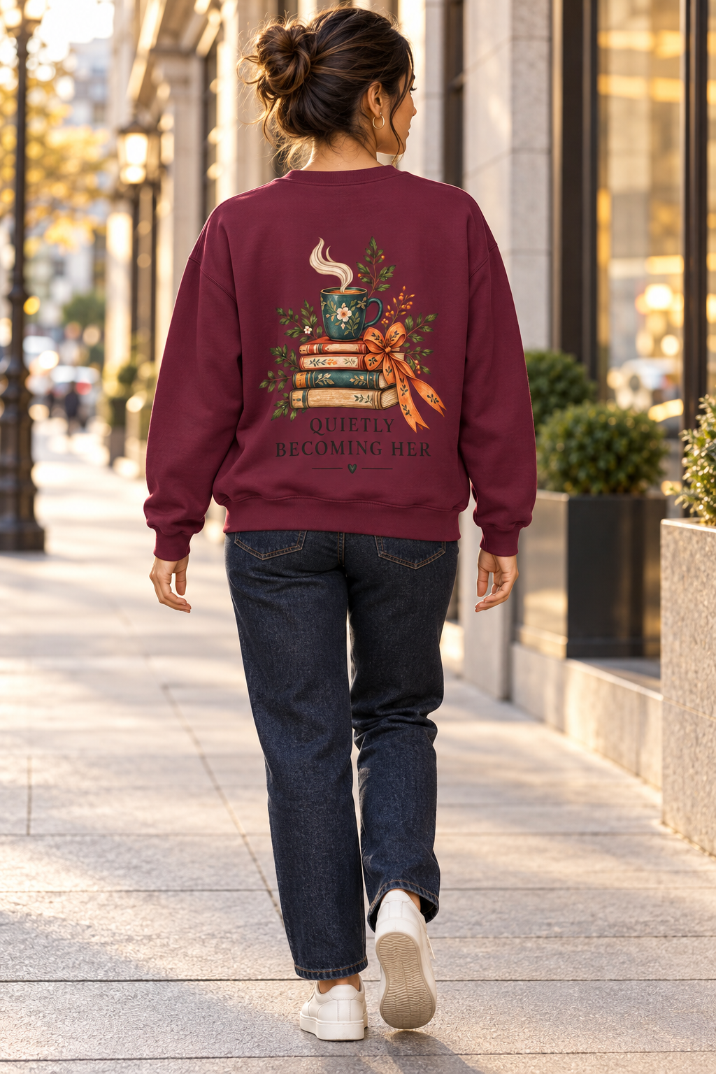 A woman viewed from behind walking on a city sidewalk, wearing a burgundy crewneck sweatshirt with a large graphic on the back featuring stacked books, a coffee mug, autumn leaves, and the text 'IT'S NOT ALL BECOMING EASY'.