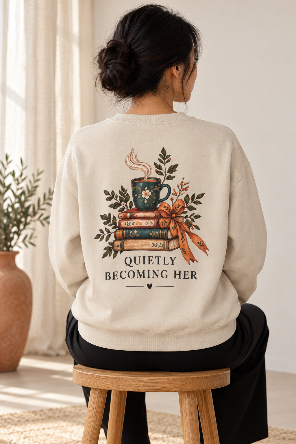 A woman viewed from behind wearing a beige sweatshirt with a large graphic on the back featuring a stack of books, a teal mug with steam,  and the text 'QUIETLY BECOMING HER'. She is sitting on a wooden stool in a bright, neutral-toned room.