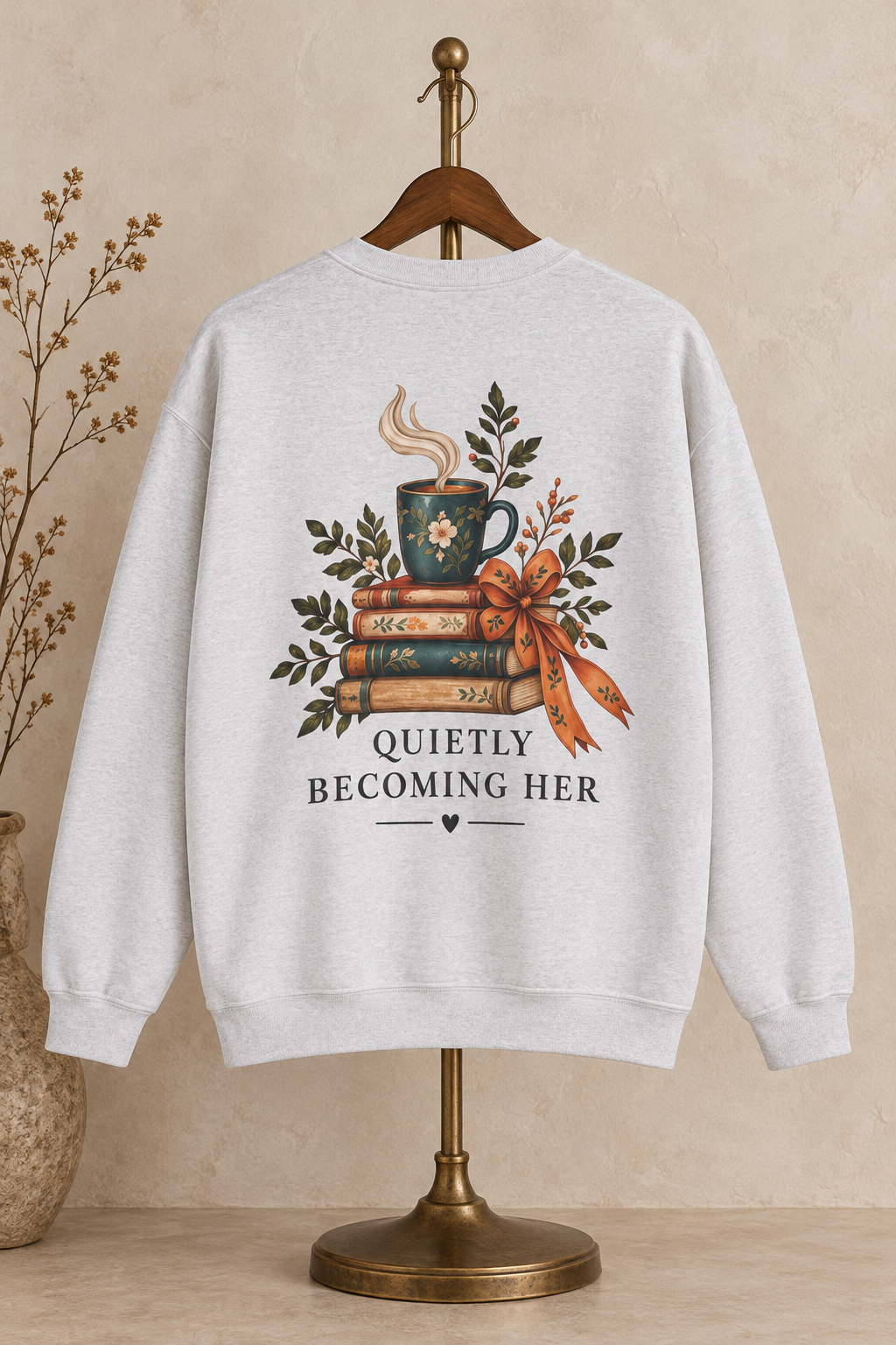 A light gray heathered crewneck sweatshirt displayed on a wooden hanger and stand, featuring a graphic of a mug on stacked books with a ribbon and the text 'QUIETLY BECOMING HER'.