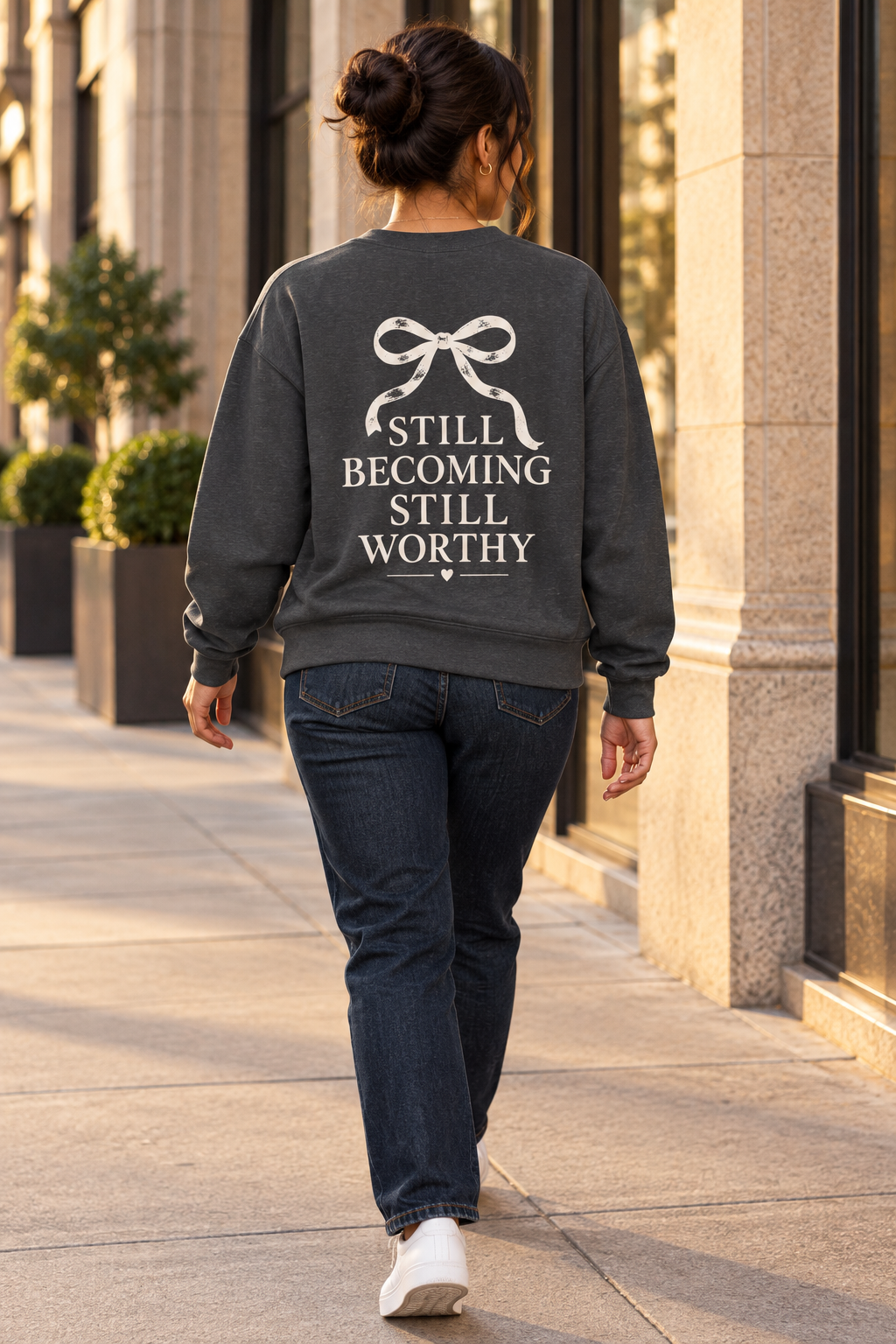 A woman viewed from behind walking on a city sidewalk, wearing a dark charcoal gray sweatshirt with a cream graphic print that reads 'STILL BECOMING STILL WORTHY' beneath a bow illustration, paired with dark blue jeans and white sneakers.