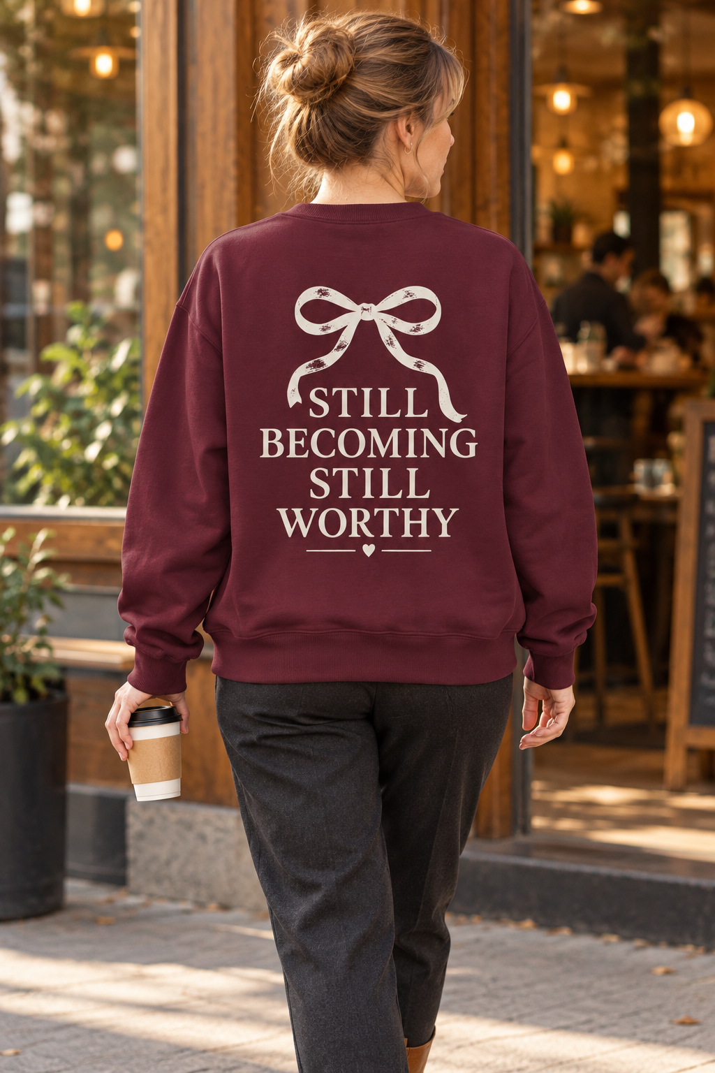 A woman viewed from behind wearing a maroon crewneck sweatshirt with a cream bow graphic and the text 'STILL BECOMING STILL WORTHY' printed on the back. She is walking outdoors on a sidewalk in front of a coffee shop, holding a takeaway coffee cup.