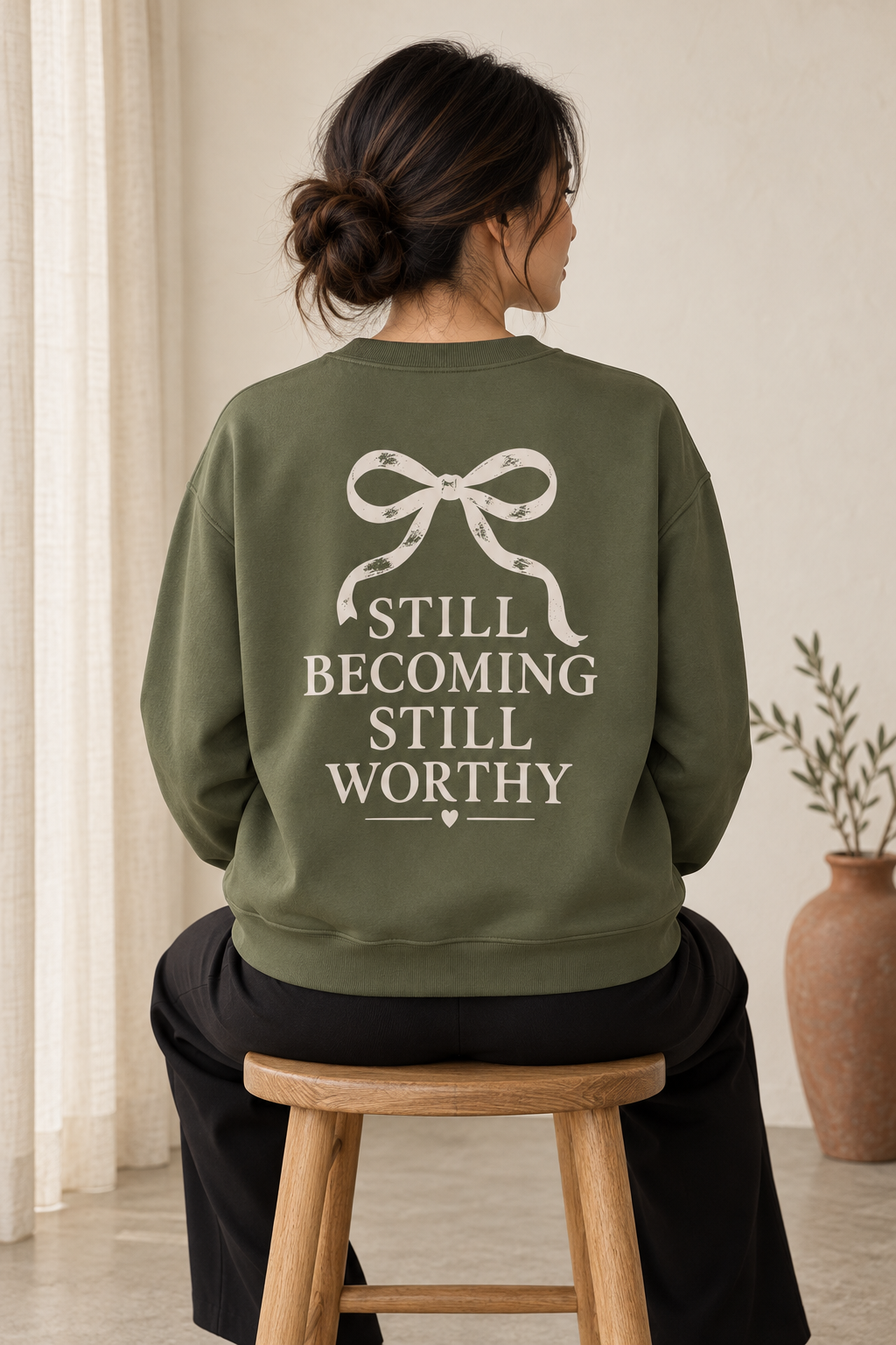 A woman seated on a wooden stool, shown from the back, wearing an olive green sweatshirt with a cream ribbon-bow graphic and the text 'STILL BECOMING STILL WORTHY' printed across the back.