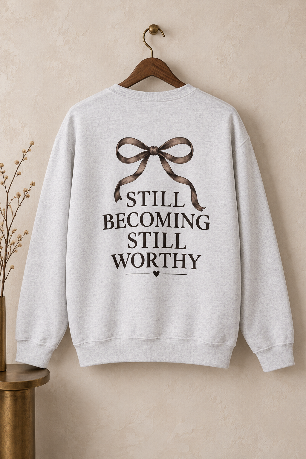 A light heather gray crewneck sweatshirt hanging on a wooden hanger against a textured beige wall. The back of the sweatshirt features a graphic of a brown ribbon bow above the dark text 'STILL BECOMING STILL WORTHY'.