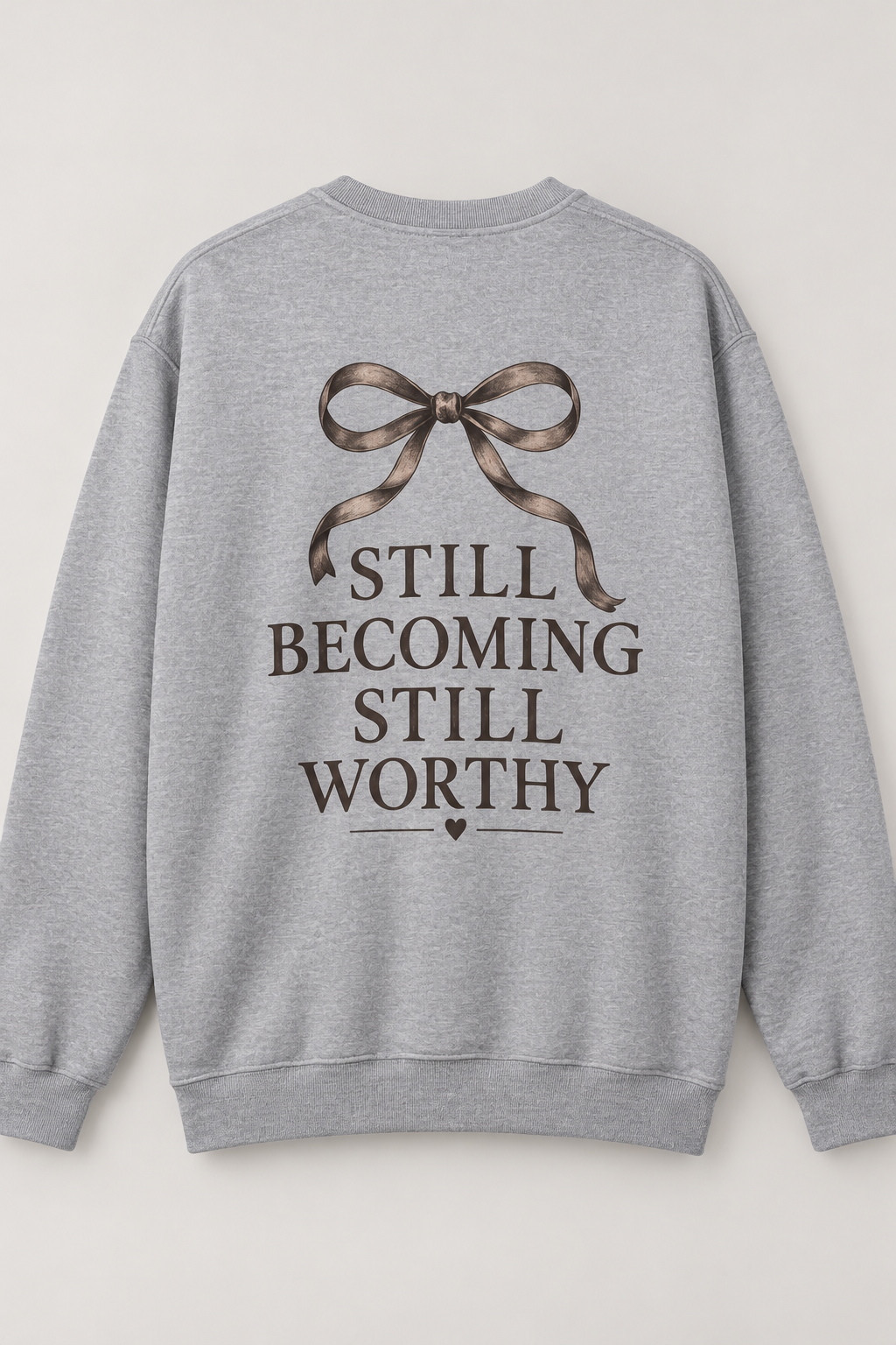 A light gray crewneck sweatshirt shown from the back, featuring a graphic print of a brown ribbon bow above the text 'STILL BECOMING STILL WORTHY' in dark brown lettering.