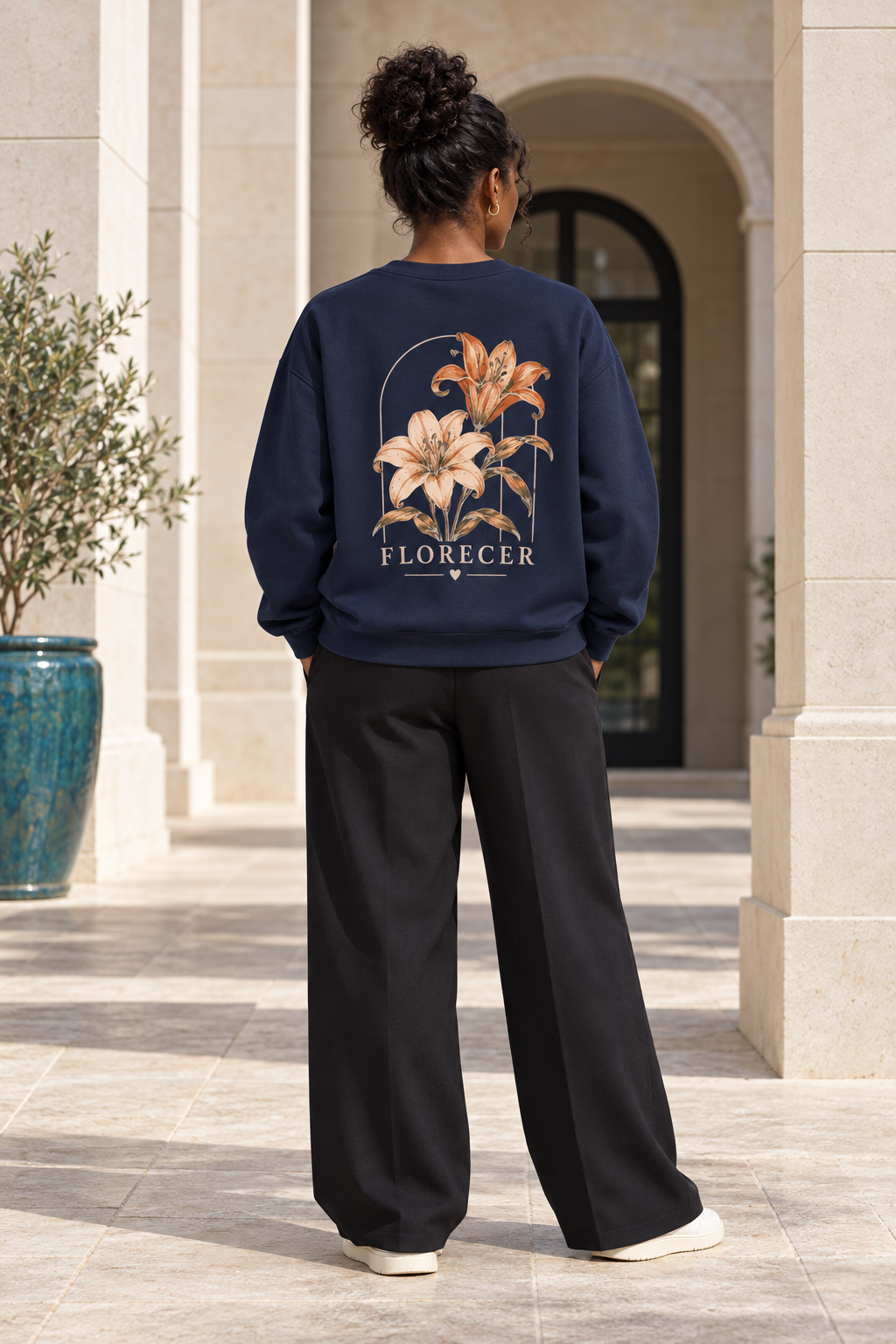 A woman stands with her back to the camera in a sunlit outdoor corridor, wearing a navy blue sweatshirt with a large floral back graphic and the word 'FLORECER' printed below it, paired with black wide-leg pants and white sneakers.