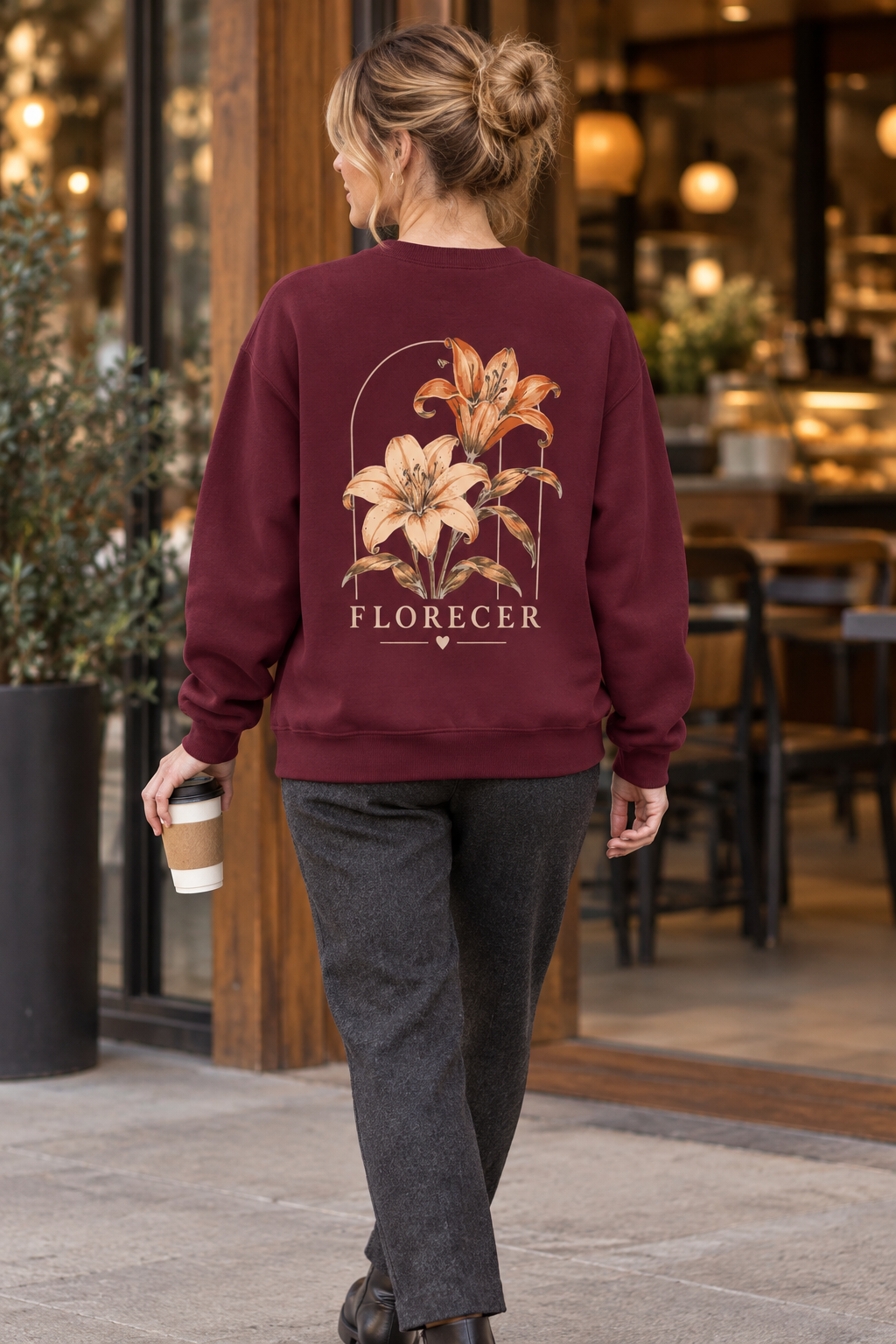 A woman viewed from behind wearing a burgundy crewneck sweatshirt with a large floral graphic and the word 'FLORECER' on the back, paired with dark gray pants, walking in front of a cafe.