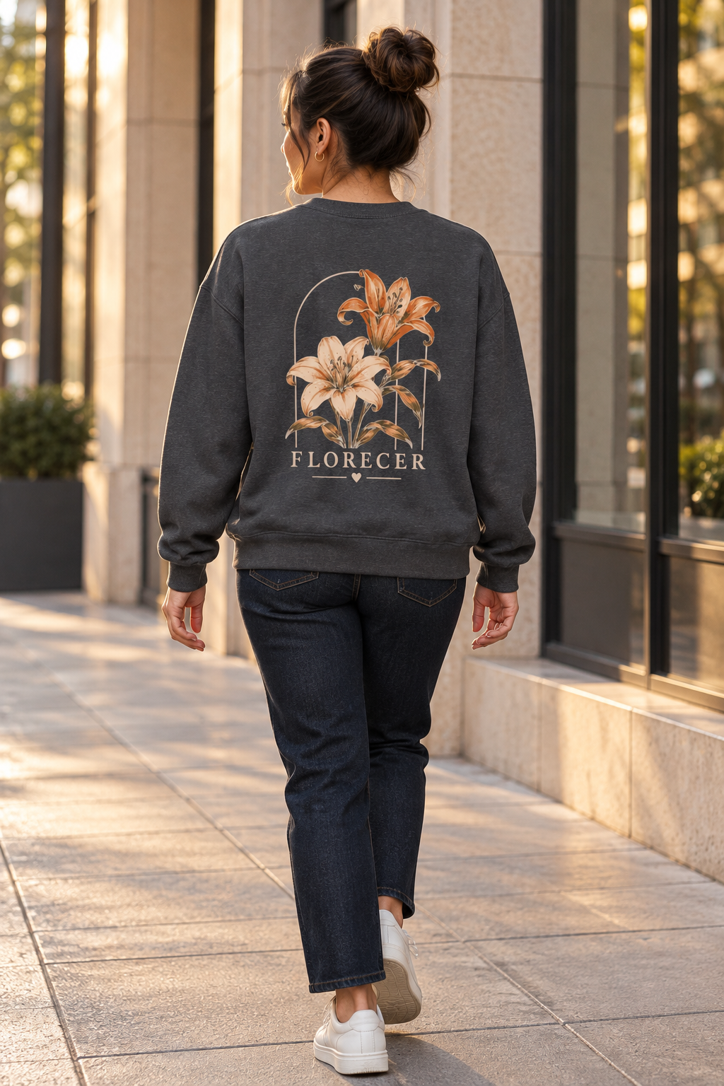 A woman viewed from behind walking on a sunlit city sidewalk, wearing a dark charcoal gray sweatshirt with a large floral graphic and the word 'FLORECER' on the back, paired with dark jeans and white sneakers.