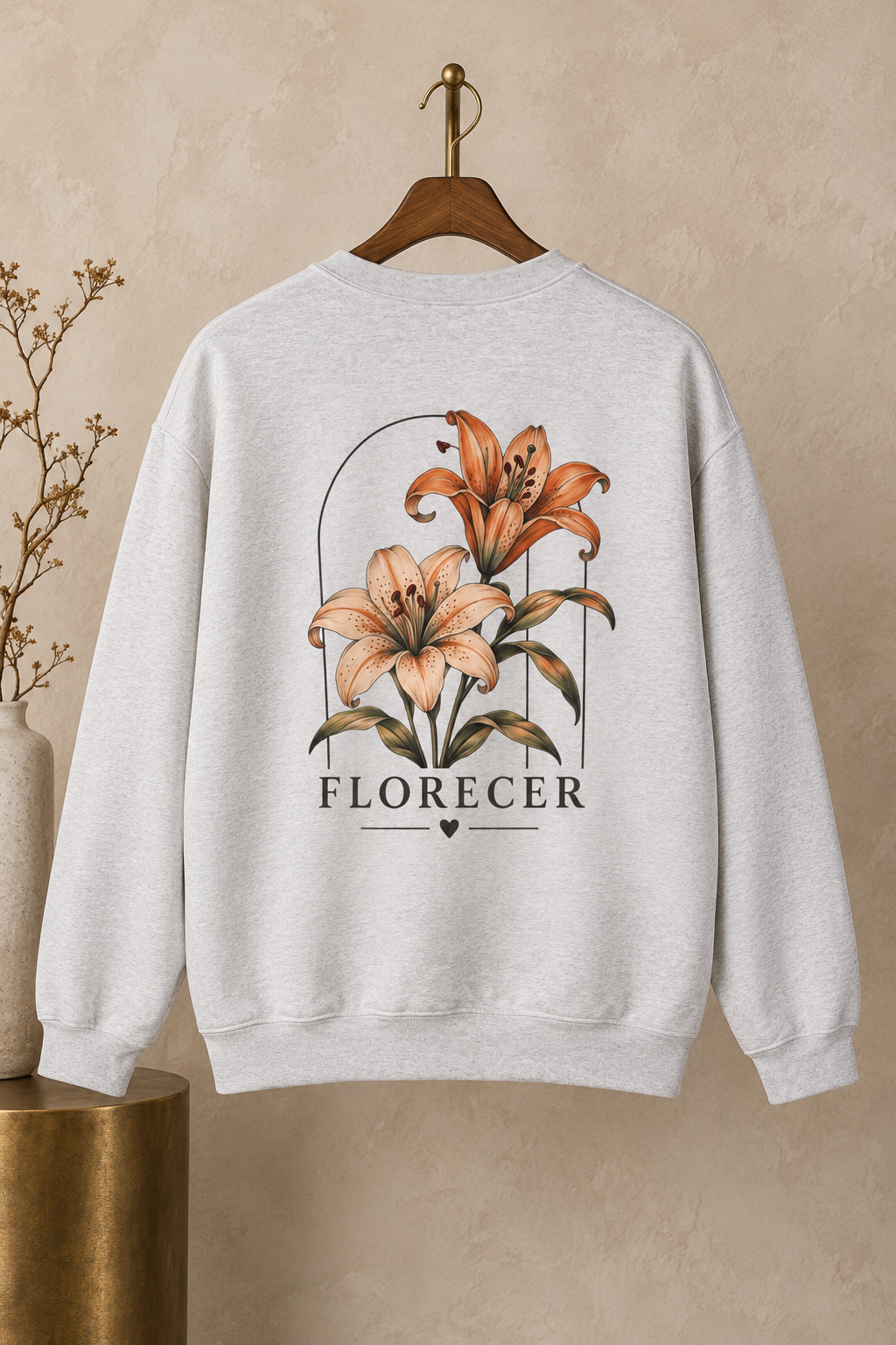 A light heather gray crewneck sweatshirt hanging on a wooden hanger against a textured beige wall. The back of the sweatshirt features a graphic print of two lilies under a minimalist arch, with the word 'FLORECER' printed below in a serif font.