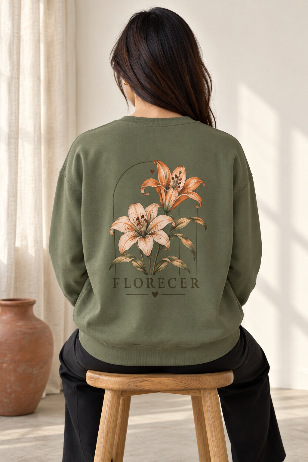 A woman viewed from behind wearing an olive green sweatshirt with a large floral back graphic and the word 'FLORECER' printed below, seated on a wooden stool in a softly lit indoor setting.