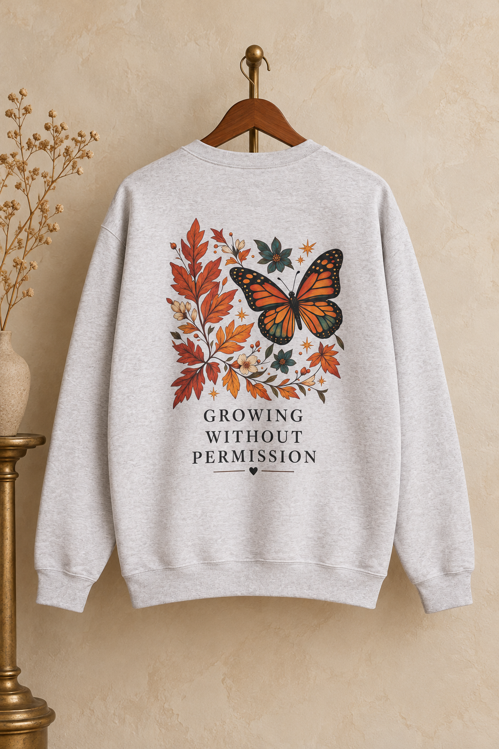 A light heather gray crewneck sweatshirt hanging on a wooden hanger against a textured beige wall. The back of the sweatshirt features a graphic print of a monarch butterfly surrounded by autumn leaves in orange, red, and green, with the text 'GROWING WITHOUT PERMISSION' printed below.