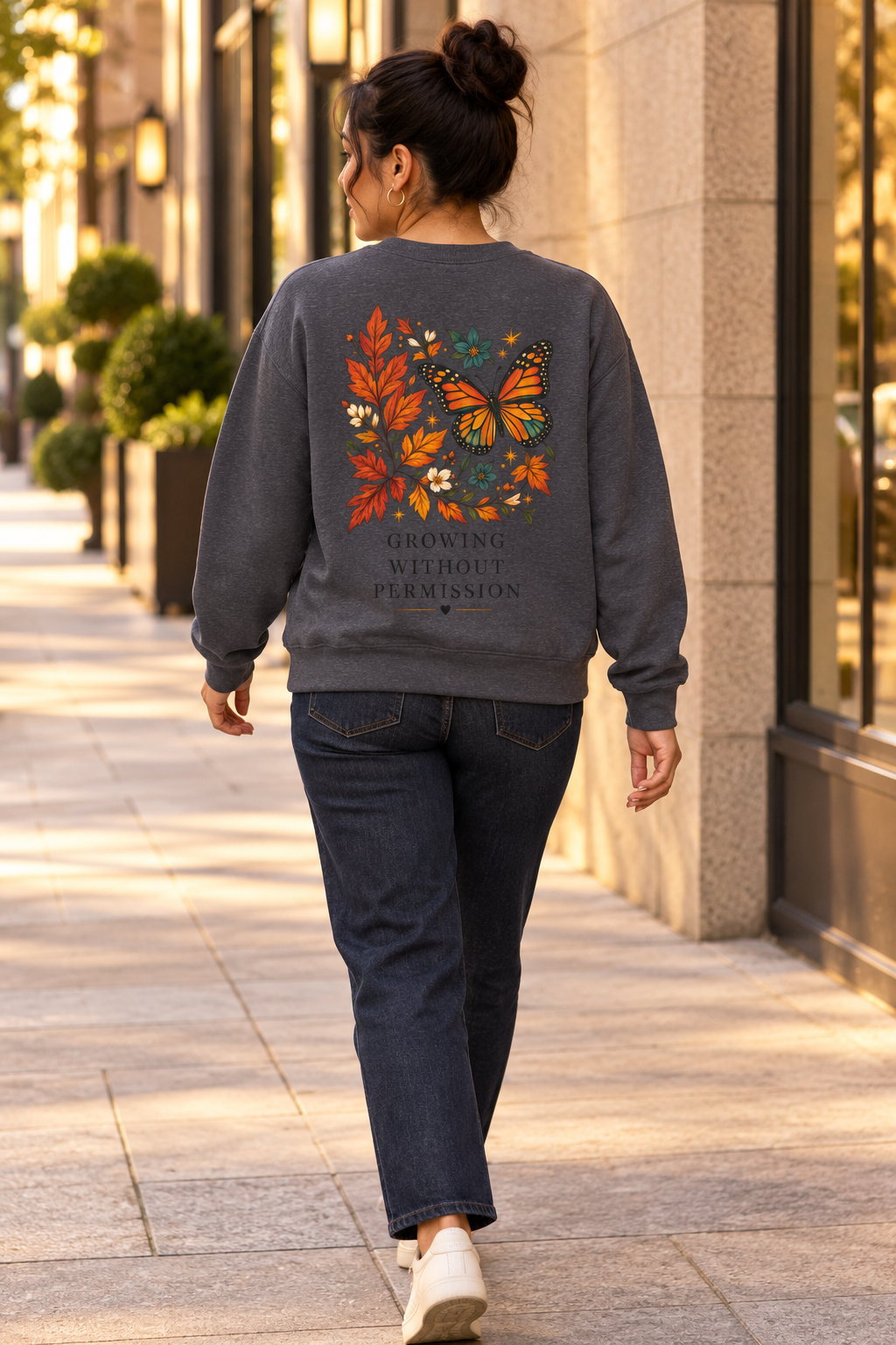 A woman viewed from behind walking on a city sidewalk, wearing a dark gray crewneck sweatshirt with a colorful butterfly and leaf graphic on the back, paired with dark blue jeans and white sneakers.